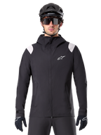 Alpinestars A-Dura Regenjacke, MTB-Regenjacke, Schwarz mit weißen Schultereinsätzen, getragen mit schwarzem MTB-Helm, Sonnenbrille und Handschuhen, wasserdichtes Design mit Kapuze und Frontreißverschluss, sportliche Outdoor-Ausrüstung für den Radsport