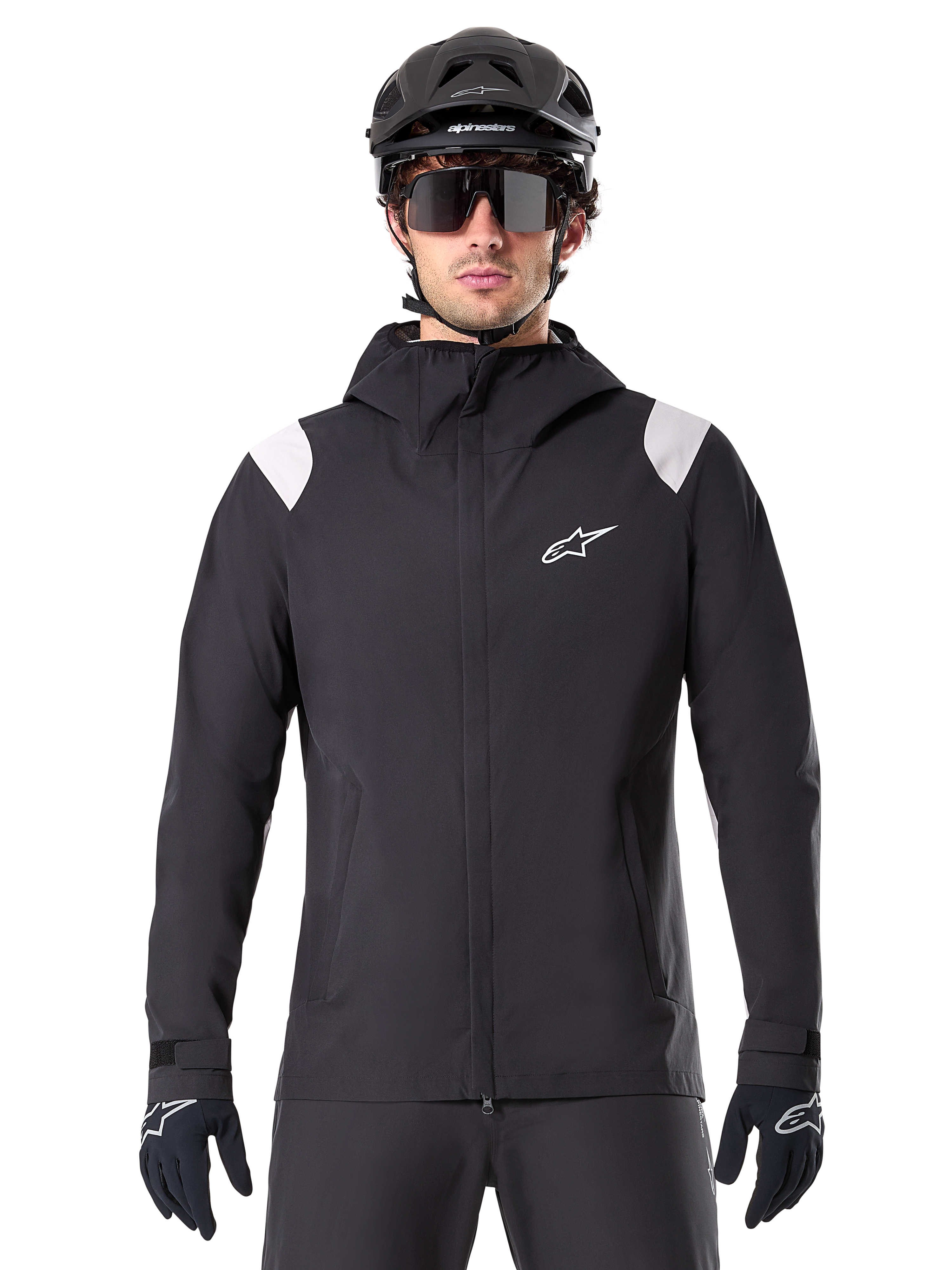 Alpinestars A-Dura Regenjacke, MTB-Regenjacke, Schwarz mit weißen Schultereinsätzen, getragen mit schwarzem MTB-Helm, Sonnenbrille und Handschuhen, wasserdichtes Design mit Kapuze und Frontreißverschluss, sportliche Outdoor-Ausrüstung für den Radsport