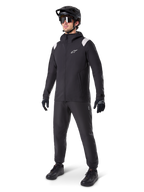 Alpinestars A-Dura Regenjacke, schwarze Mountainbike-Regenjacke mit weißen Akzenten an den Schultern, getragen von einem Fahrer mit passendem schwarzem MTB-Helm, Sonnenbrille, Handschuhen und Hose, technisches Allwetter-Radsport-Outfit