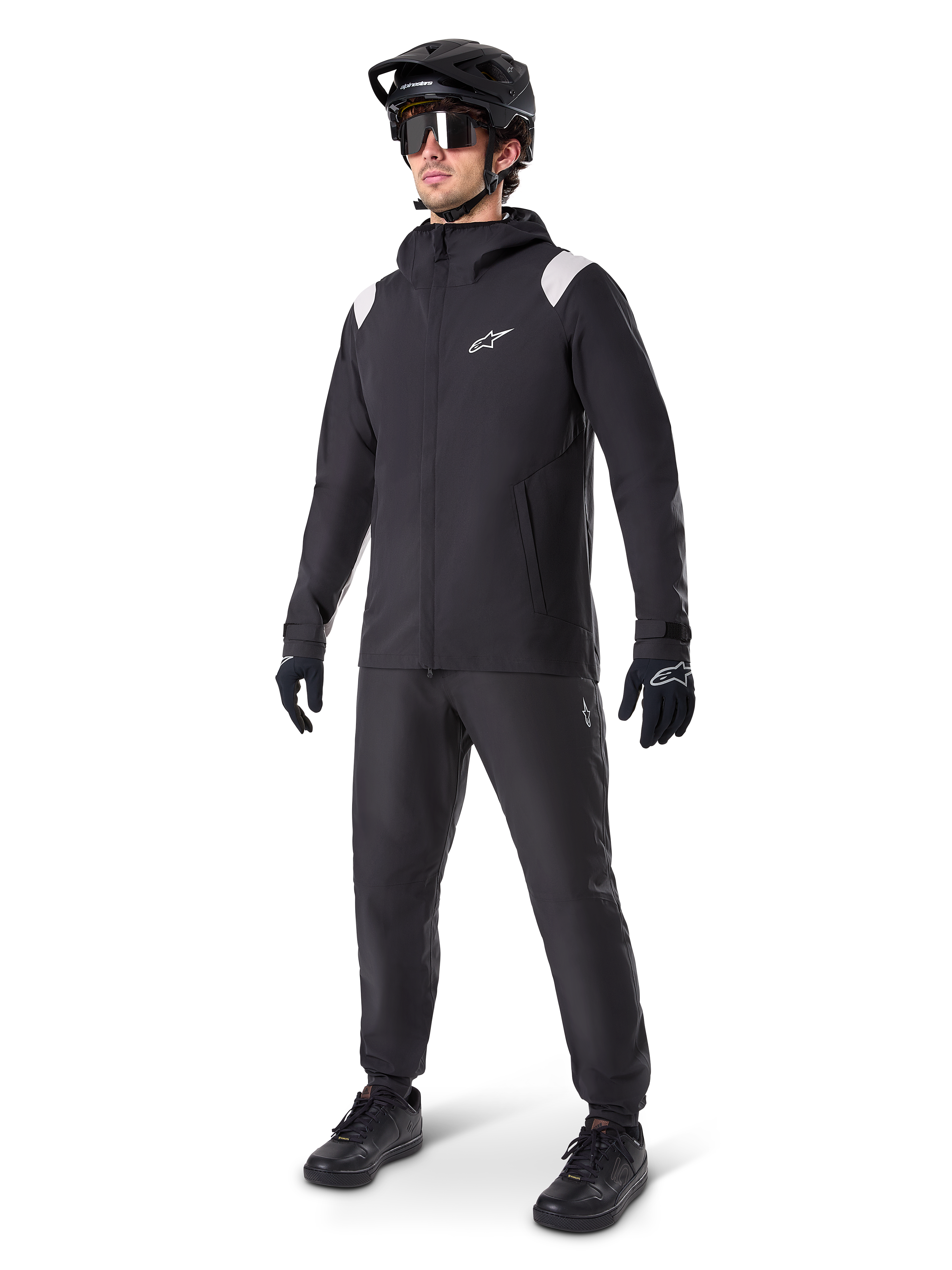 Alpinestars A-Dura Regenjacke, schwarze Mountainbike-Regenjacke mit weißen Akzenten an den Schultern, getragen von einem Fahrer mit passendem schwarzem MTB-Helm, Sonnenbrille, Handschuhen und Hose, technisches Allwetter-Radsport-Outfit