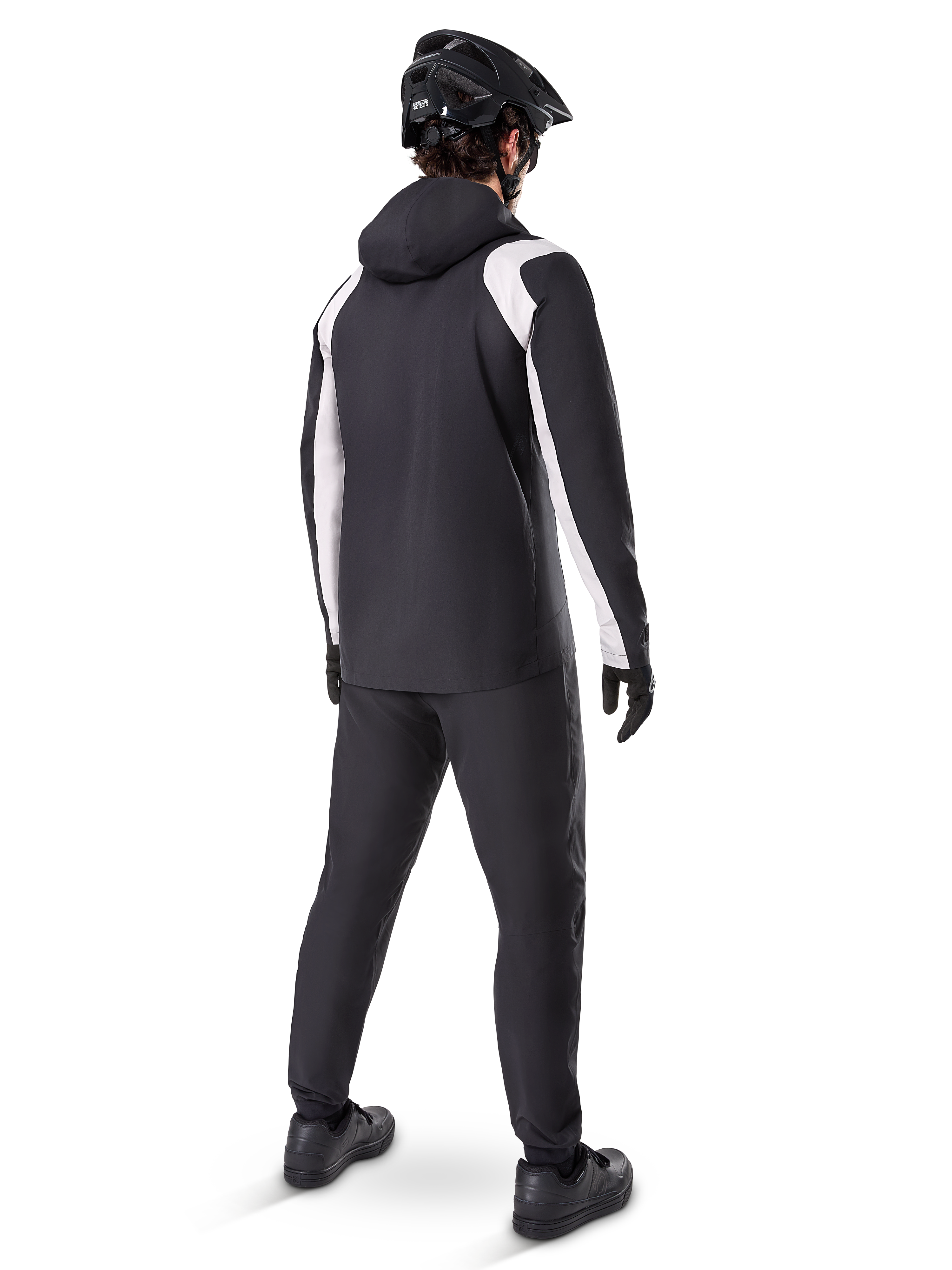Alpinestars A-Dura Regenjacke, Rückansicht eines männlichen Radfahrers in der schwarz-weißen wasserdichten MTB-Jacke, dazu passende schwarze Radhose, schwarzer Helm und Handschuhe, technische Ausrüstung für das Mountainbiken bei nassen Bedingungen.