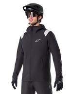 Alpinestars A-Dura Regenjacke, Mountainbike-Regenjacke, Schwarz mit hellgrauen Schulterakzenten, getragen von einem Fahrer mit schwarzem MTB-Helm und Sonnenbrille, weißes Logo auf der Brust, wasserdichte Konstruktion, kombiniert mit schwarzer Hose und Handschuhen für den Offroad-Radsport