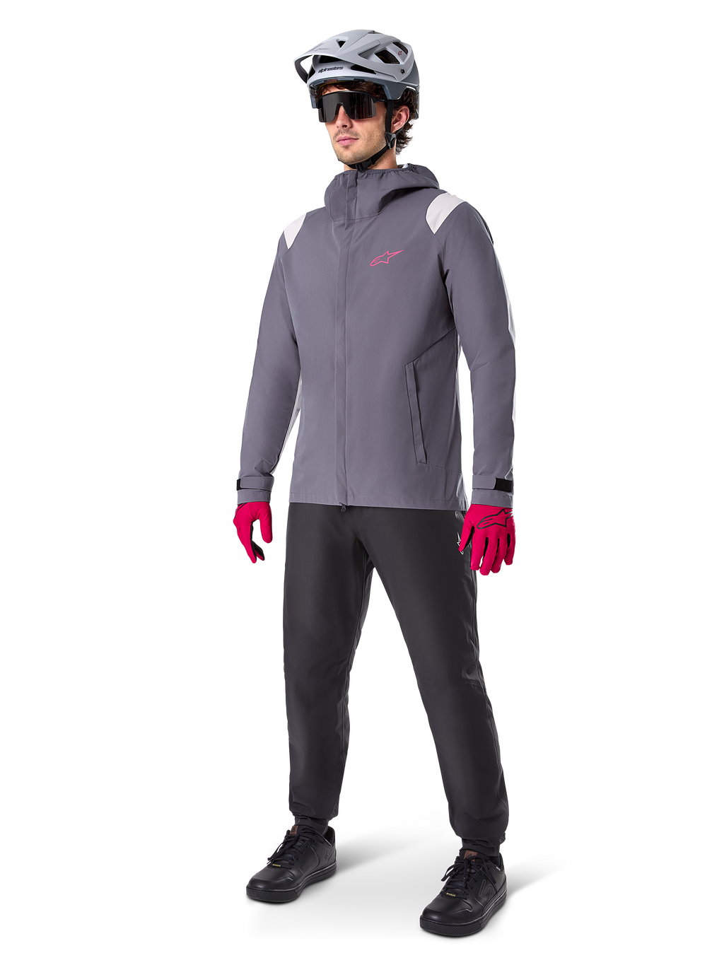 Männliches Model in der Alpinestars A-Dura Regenjacke in Dark Titanium, graue Kapuzenjacke mit pinkem Brustlogo und hellgrauen Schultereinsätzen, kombiniert mit einem grauen MTB-Helm, dunkler Sonnenbrille, leuchtend pinken Handschuhen und einer schwarzen Hose für den Offroad-Radsport.