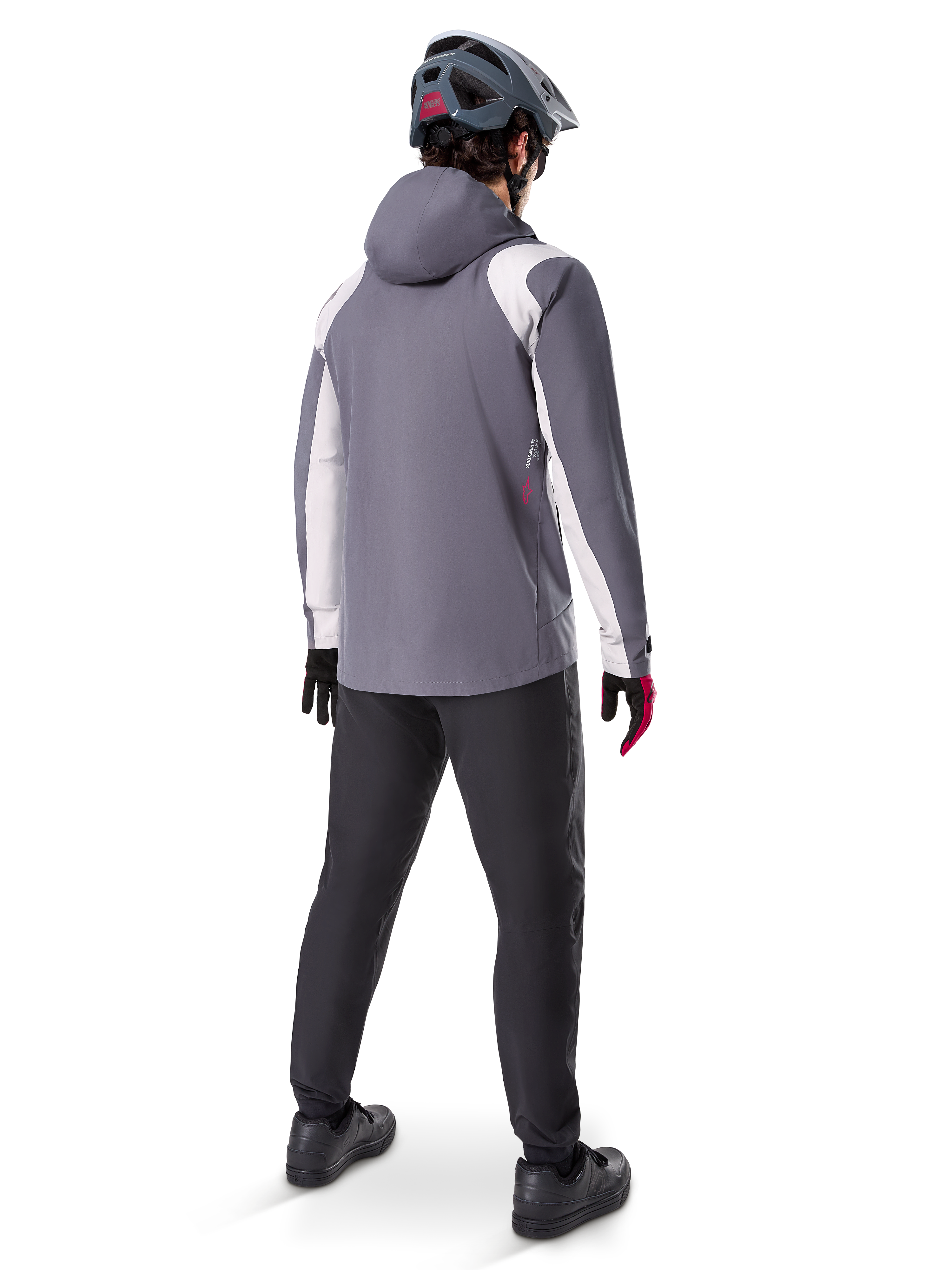 Alpinestars A-Dura Regenjacke, Mountainbike-Jacke, Dunkles Titangrau mit weißen Einsätzen, Rückansicht eines Fahrers mit passendem grauen Helm und schwarzer Hose, technische Outdoor-Ausrüstung für nasses Wetter