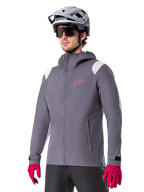 Alpinestars A-Dura Regenjacke, Mountainbike-Regenjacke, Dark Titanium Grau mit pinkem Logo und weißen Schulterakzenten, Kapuzendesign mit Fronttaschen, getragen mit pinken Handschuhen und einem hellgrauen Fahrradhelm