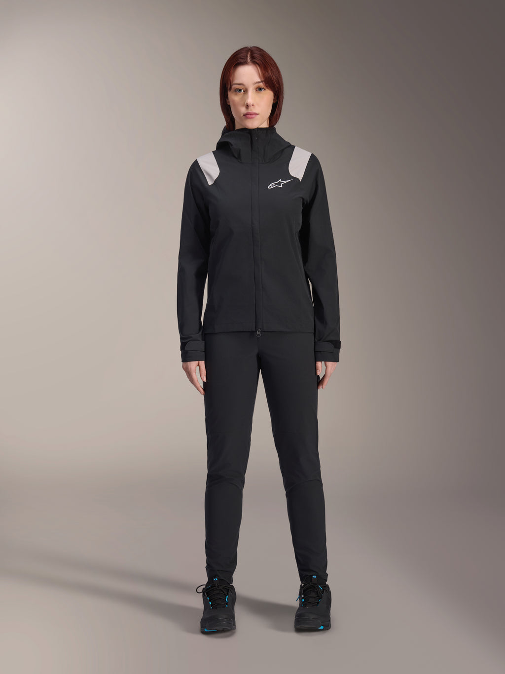 Alpinestars Stella A-Dura Regenjacke, Damen MTB-Regenjacke, Schwarz mit hellgrauen Schulterakzenten und weißem Logo, getragen mit passender schwarzer Hose und Schuhen, mit Kapuze und verstellbaren Manschetten, entwickelt für Mountainbike-Trailfahrten