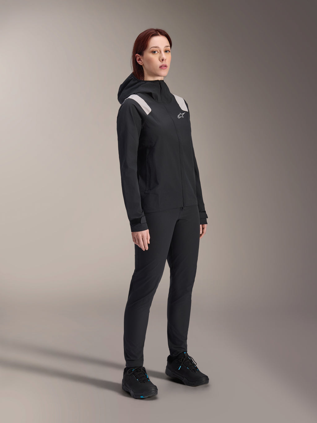 Frau in der Alpinestars Stella A-Dura Regenjacke in Schwarz, 2,5-lagiges wasserdichtes MTB-Shell mit hellgrauen Schulterakzenten, passender schwarzer Hose und Fahrradschuhen, Studioporträt für Performance-Radsportbekleidung