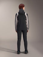 Alpinestars Stella A-Dura Regenjacke für Damen, Rückansicht einer Fahrerin, Schwarz mit hellgrauen Einsätzen an Ärmeln und Schultern, aufgesetzte Kapuze, kombiniert mit schwarzer Hose und Fahrradschuhen, technische Mountainbike-Regenausrüstung