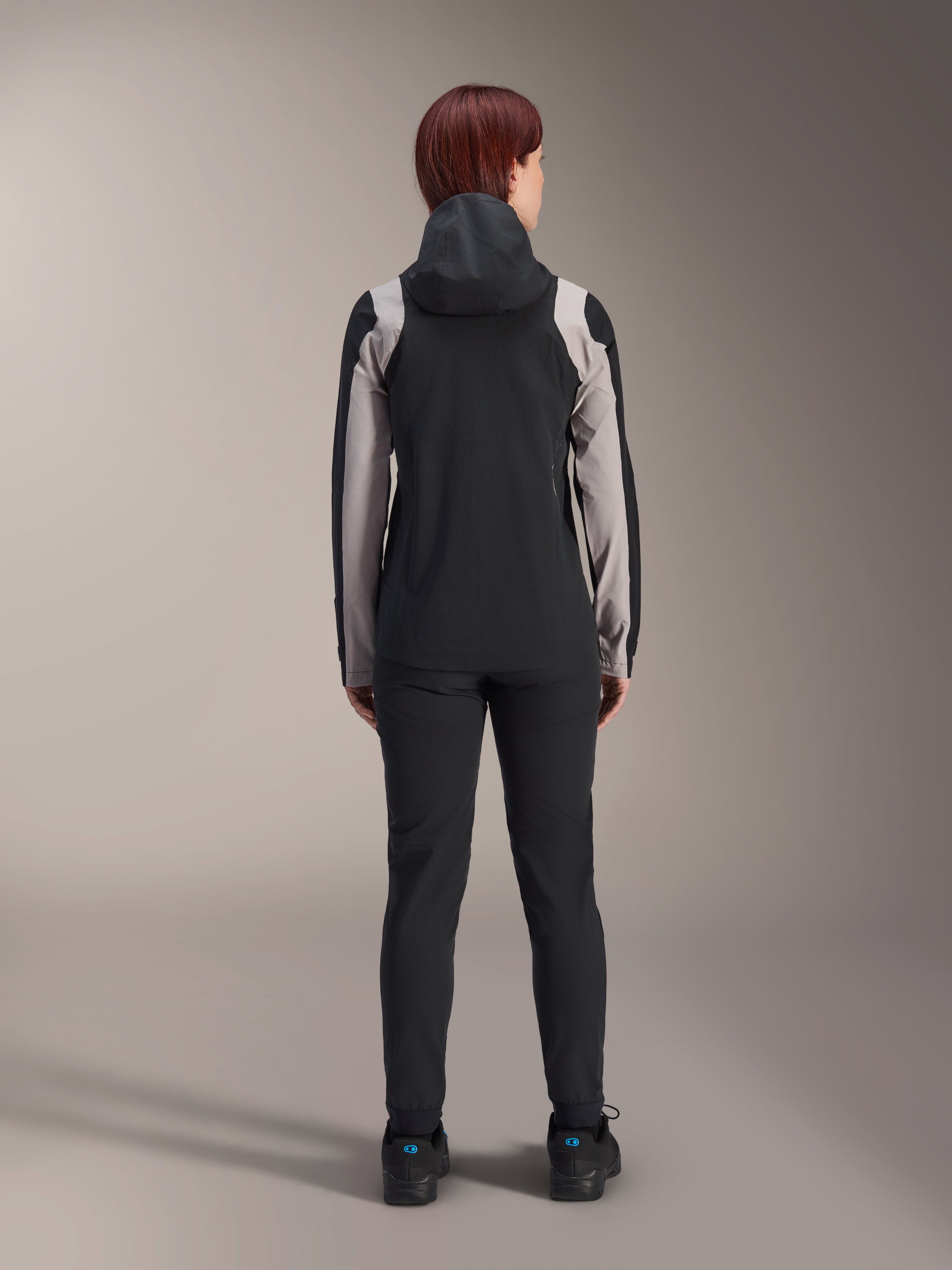 Alpinestars Stella A-Dura Regenjacke für Damen, Rückansicht einer Fahrerin, Schwarz mit hellgrauen Einsätzen an Ärmeln und Schultern, aufgesetzte Kapuze, kombiniert mit schwarzer Hose und Fahrradschuhen, technische Mountainbike-Regenausrüstung