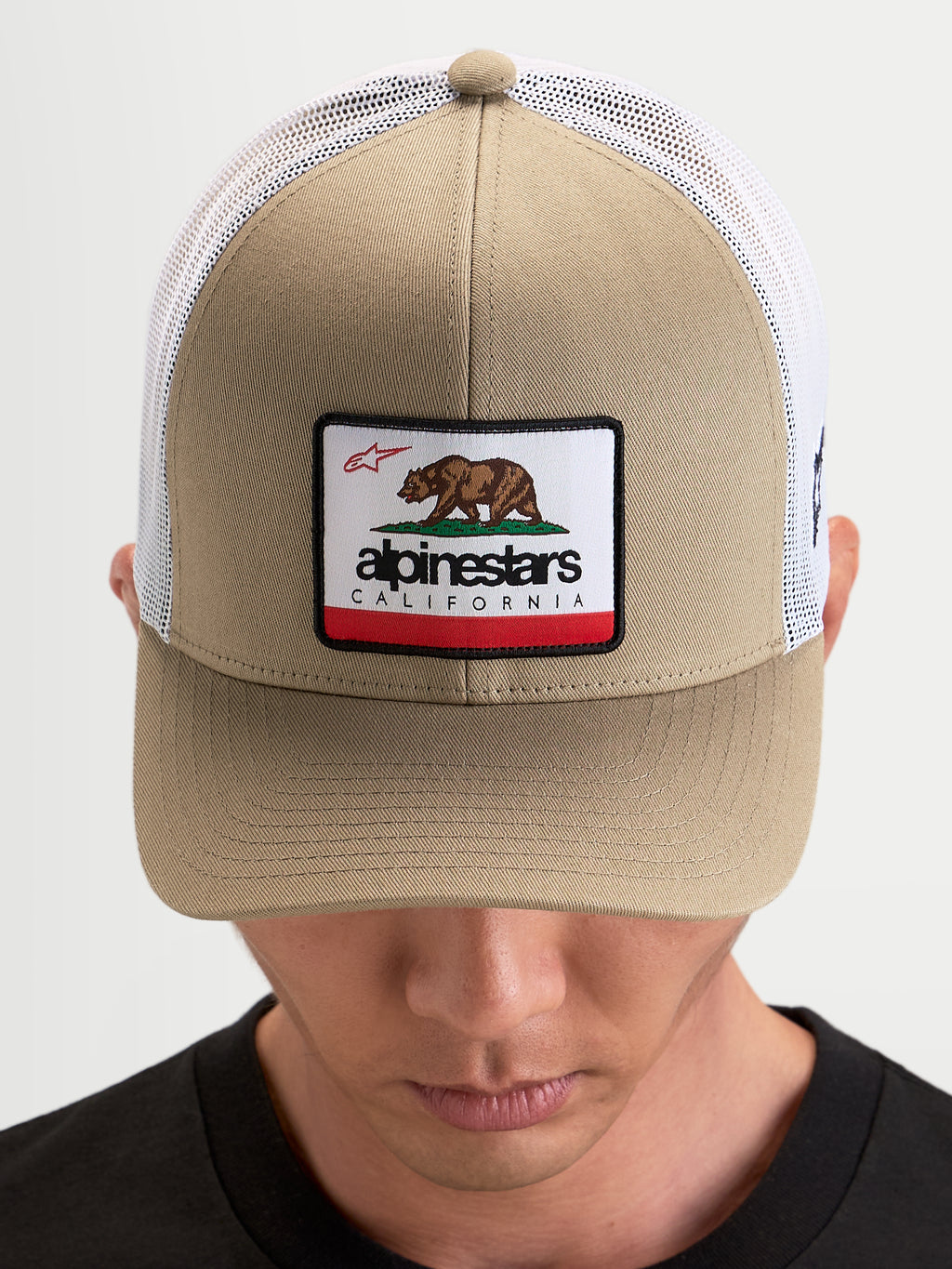 Alpinestars Cali 2.0 Kappe, Sportswear Trucker-Kappe in Khaki und Weiß, mit strukturierter Twill-Front, California-Bären-Patch, gebogenem Schirm und atmungsaktiver Mesh-Rückseite, getragen von einem Model