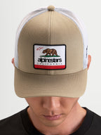 Alpinestars Cali 2.0 Kappe, Sportswear Trucker-Kappe in Khaki und Weiß, mit strukturierter Twill-Front, California-Bären-Patch, gebogenem Schirm und atmungsaktiver Mesh-Rückseite, getragen von einem Model