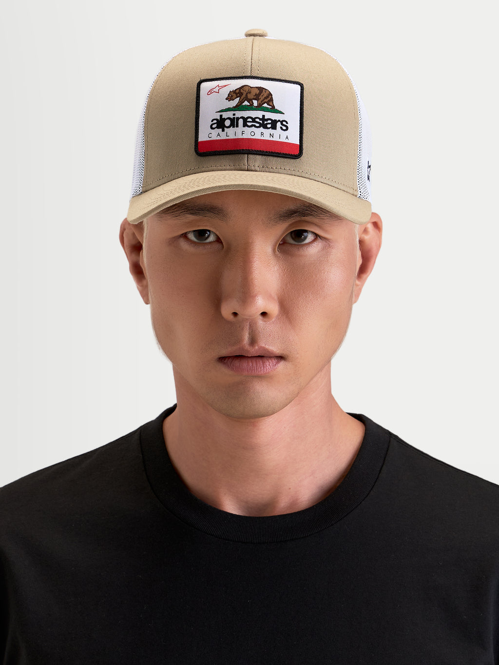 Alpinestars Cali 2.0 Kappe, Sportswear Trucker-Kappe in Khaki und Weiß, getragen von einem Model, mit gebogenem Schirm, strukturierter Twill-Front, atmungsaktiver Mesh-Rückseite und California-Bären-Patch auf der Vorderseite