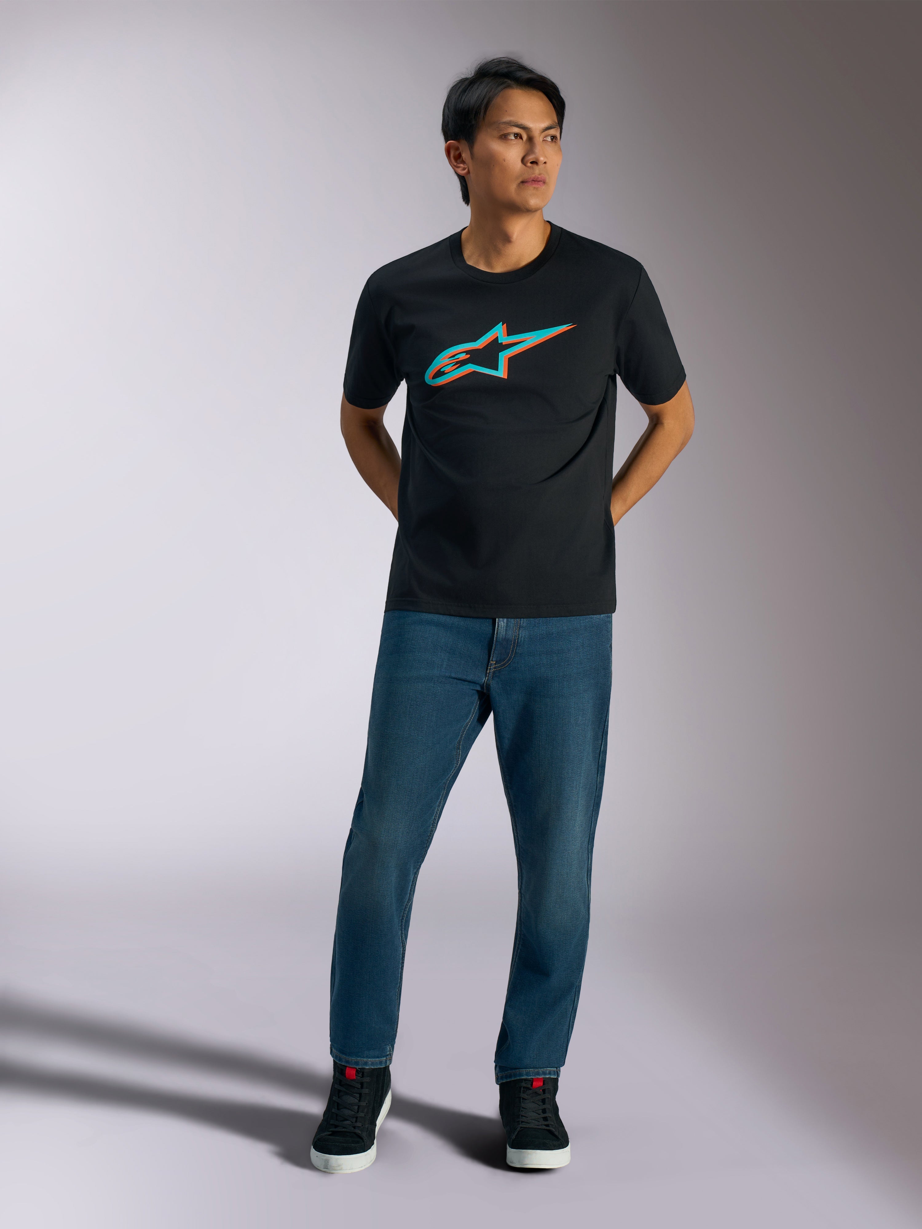 Alpinestars Ageless Shadow CSF T-Shirt, Kurzärmeliges T-Shirt, Schwarz, Türkis- und orangefarbenes Ageless Shadow Logo auf der Brust, getragen von einem Model mit blauen Jeans und schwarzen Schuhen, lässige Motorsport-Streetwear
