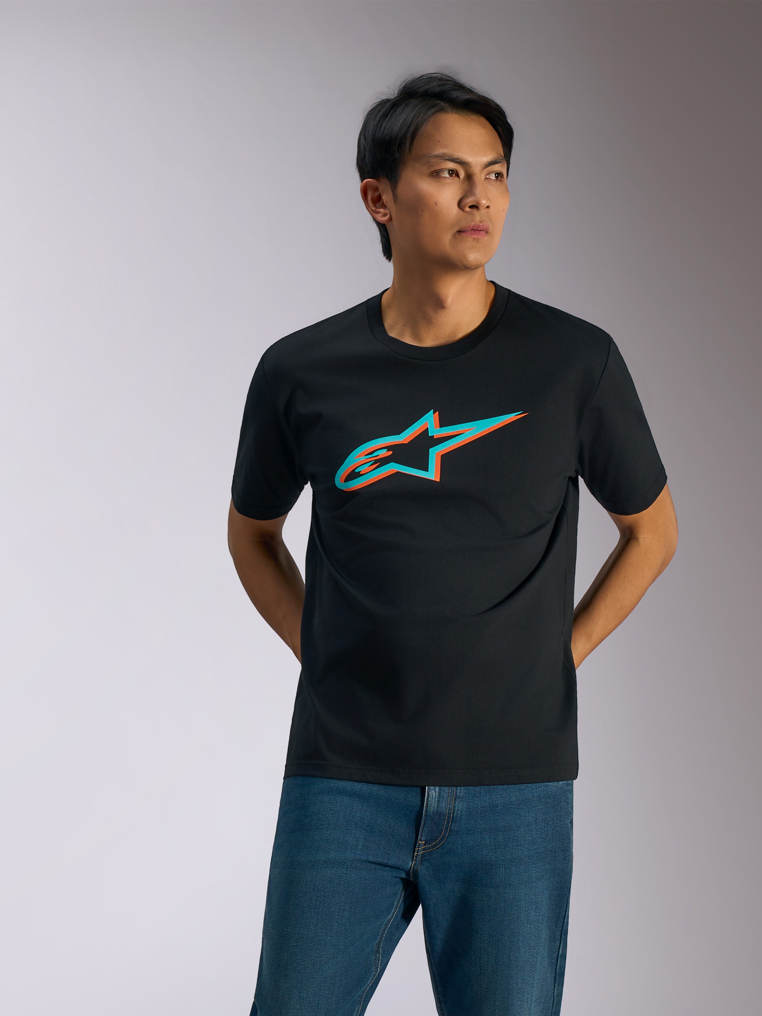 Alpinestars Ageless Shadow CSF Tee, Kurzarm-T-Shirt, Schwarz mit türkisem und orangem Shadow-Logo, getragen von einem Mann mit blauen Jeans, lässige Motorsport-Streetwear