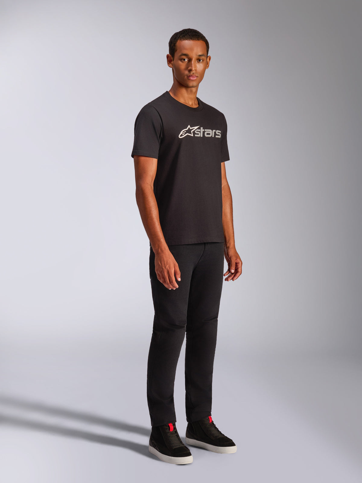 Alpinestars Blaze 2.0 CSF Tee, kurzärmeliges T-Shirt, Schwarz mit weißer und grauer Logo-Grafik, getragen von einem männlichen Model mit schwarzer Hose und High-Top-Sneakern, lässige Motorsport-Streetwear