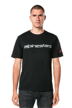Alpinestars Linear Wordmark 2.0 CSF Tee, kurzärmeliges T-Shirt, Schwarz, graue Linear-Wortmarke auf der Brust und rotes Ageless-Logo am Ärmel, getragen von einem Model mit dunkler Jeans, lässige vom Motorsport inspirierte Streetwear