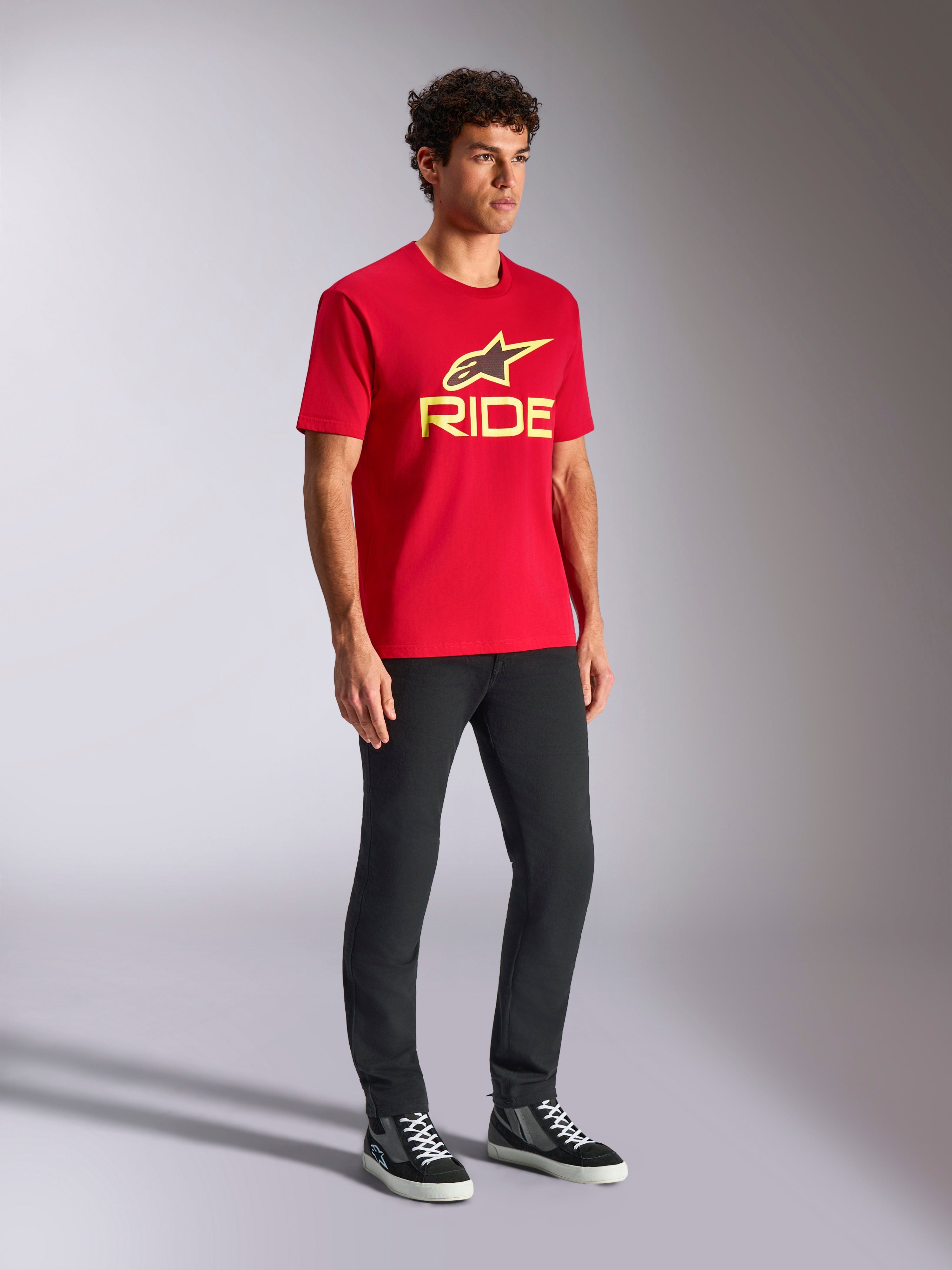 Alpinestars Ride 4.0 CSF T-Shirt, kurzärmeliges T-Shirt, Rot mit gelbem und schwarzem Logo auf der Vorderseite, kombiniert mit schwarzer Hose und Sneakern, lässige Motorsport-Streetwear