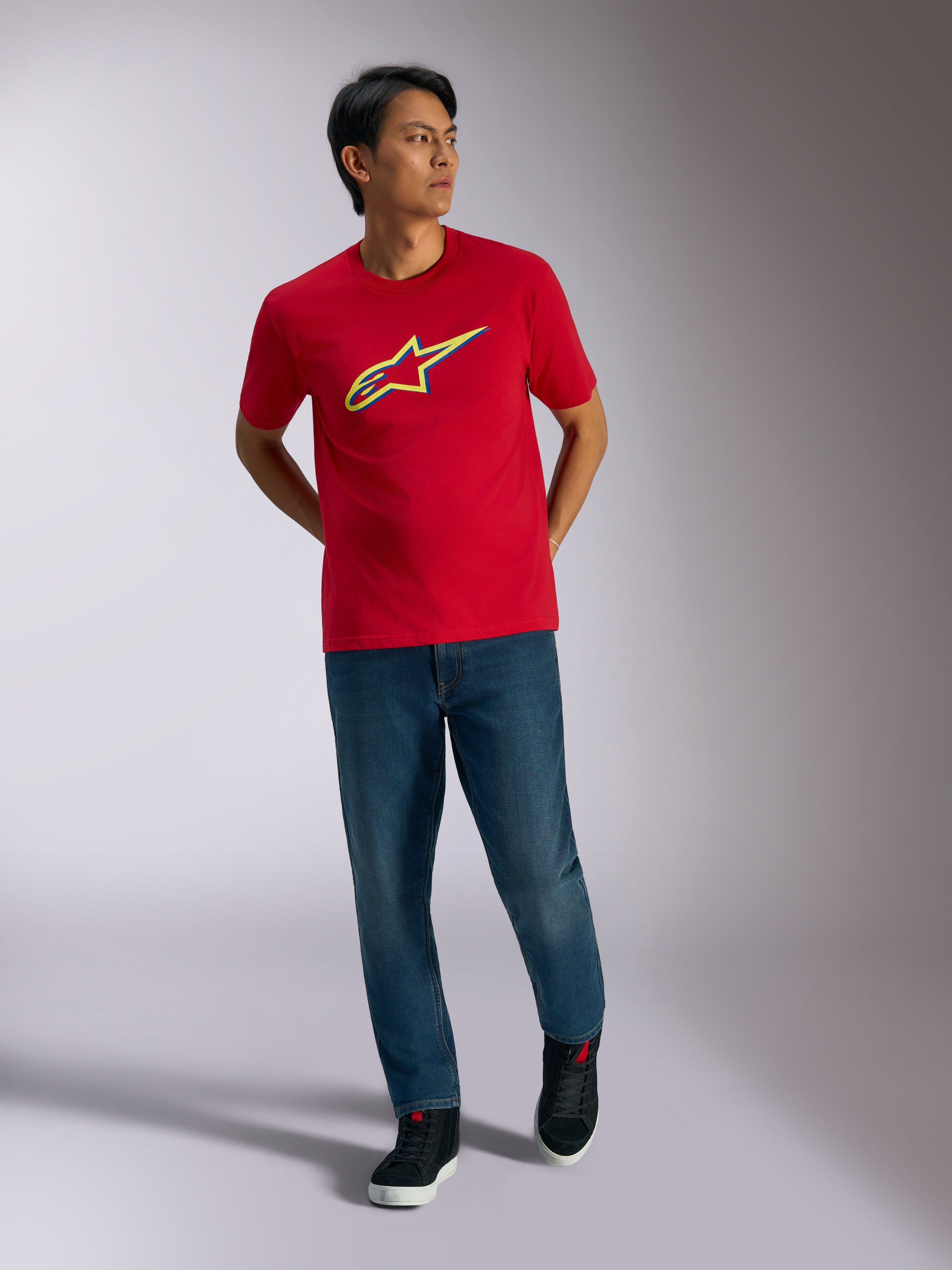 Alpinestars Ageless Shadow CSF T-Shirt, rotes Kurzarm-T-Shirt, gelbe und blaue Schattenlogo-Grafik, getragen mit blauen Jeans und schwarzen Sneakern, lässige Motorsport-Lifestyle-Bekleidung