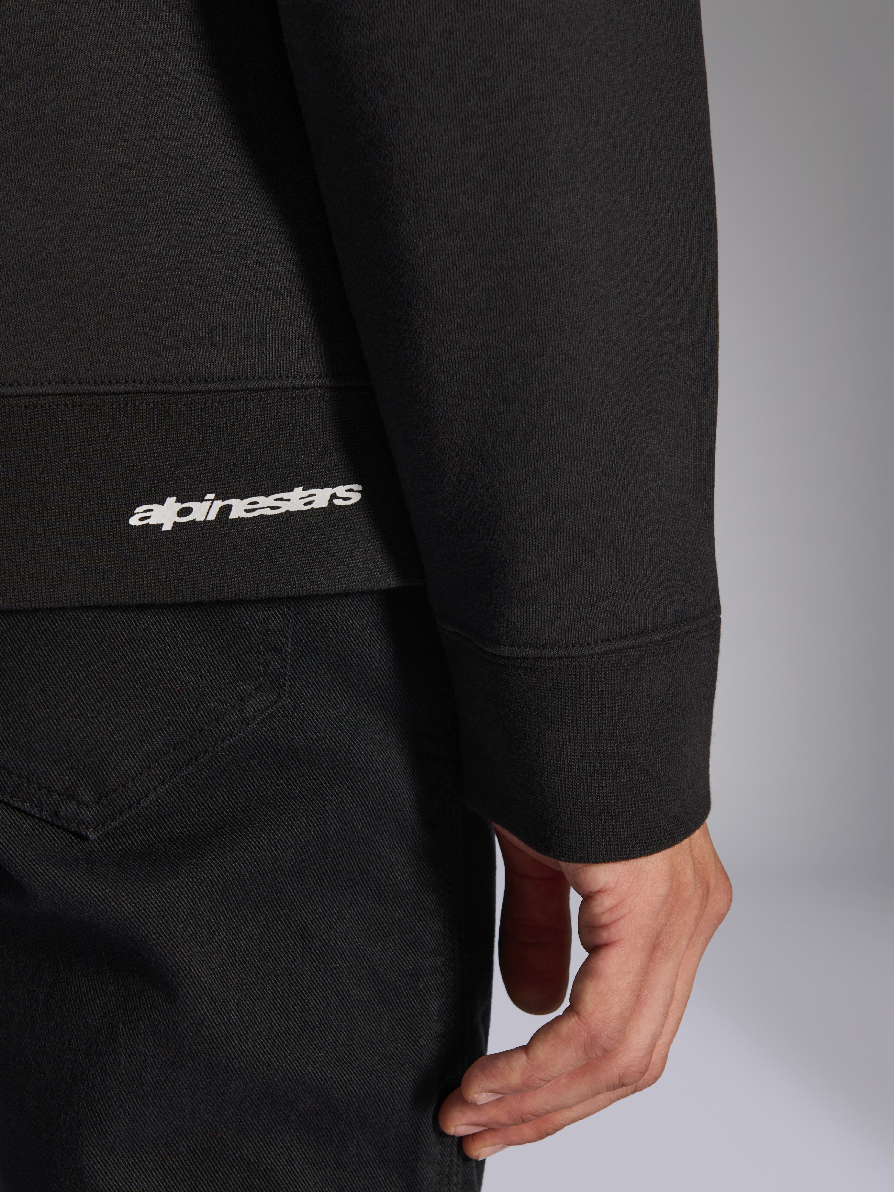 Alpinestars Positive Crew, schwarzes Pullover-Sweatshirt, Detailaufnahme von geripptem Saum und Ärmelbündchen mit weißem Alpinestars-Logo, hochwertige Stofftextur, lässige vom Motorsport inspirierte Sportbekleidung