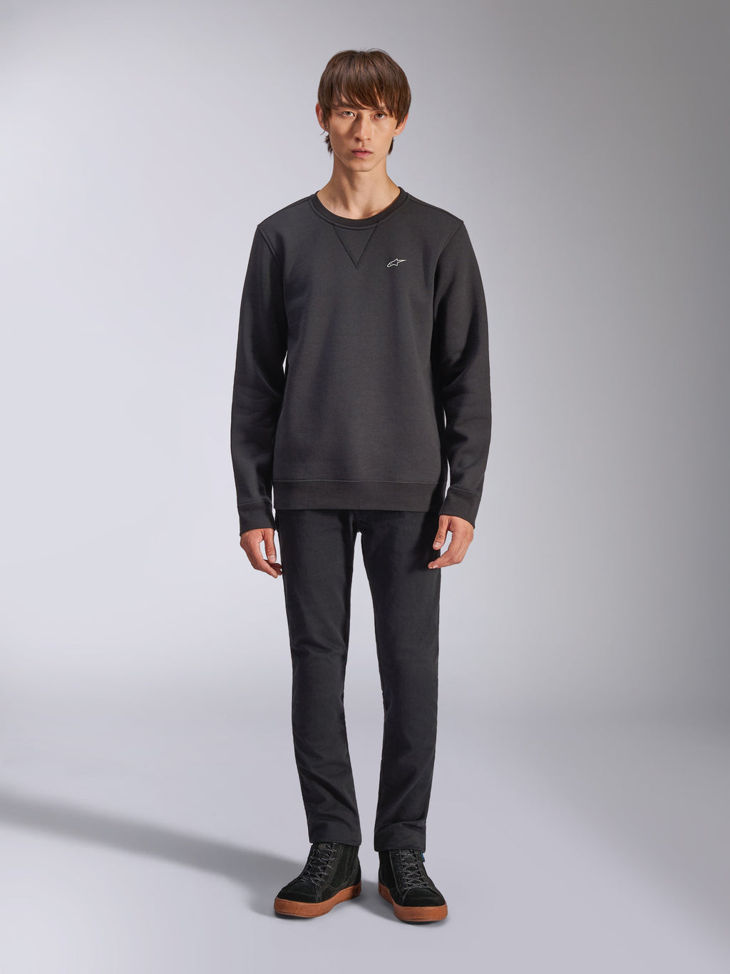 Alpinestars Positive Crew Sweatshirt, lässiger schwarzer Crewneck, kleines weißes Logo auf der Brust, V-Stitch-Ausschnitt, Ganzkörper-Studioaufnahme mit Model in schwarzer Hose und Sneakern, Motorsport-inspirierte Lifestyle-Bekleidung