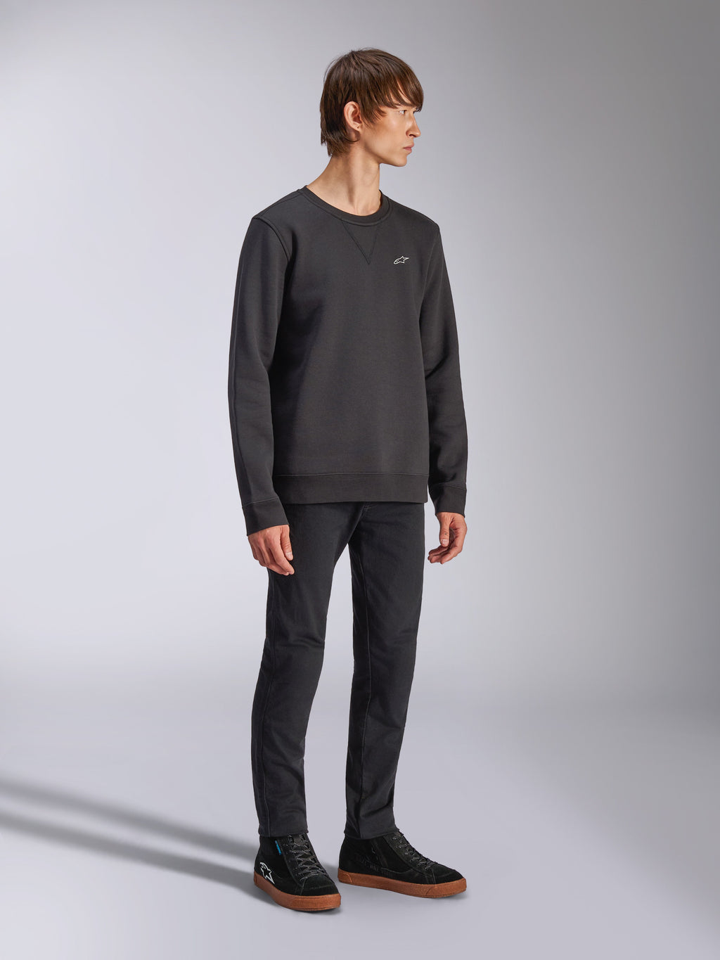 Alpinestars Positive Crew, Pullover-Sweatshirt, Schwarz, kleines weißes Alpinestars-Logo auf der Brust, gerippter Kragen mit V-Detail, Rippbündchen an Ärmeln und Saum, getragen von einem Model mit schwarzer Hose und Sneakern, lässige vom Motorsport inspirierte Streetwear