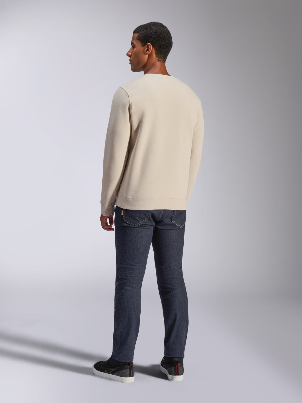 Alpinestars Positive Crew, Off-White Rundhals-Sweatshirt, Rückansicht am Modell, kombiniert mit dunklen Denim-Jeans und schwarzen Sneakern, lässige Motorsport-Streetwear, mit gerippten Bündchen und Saum