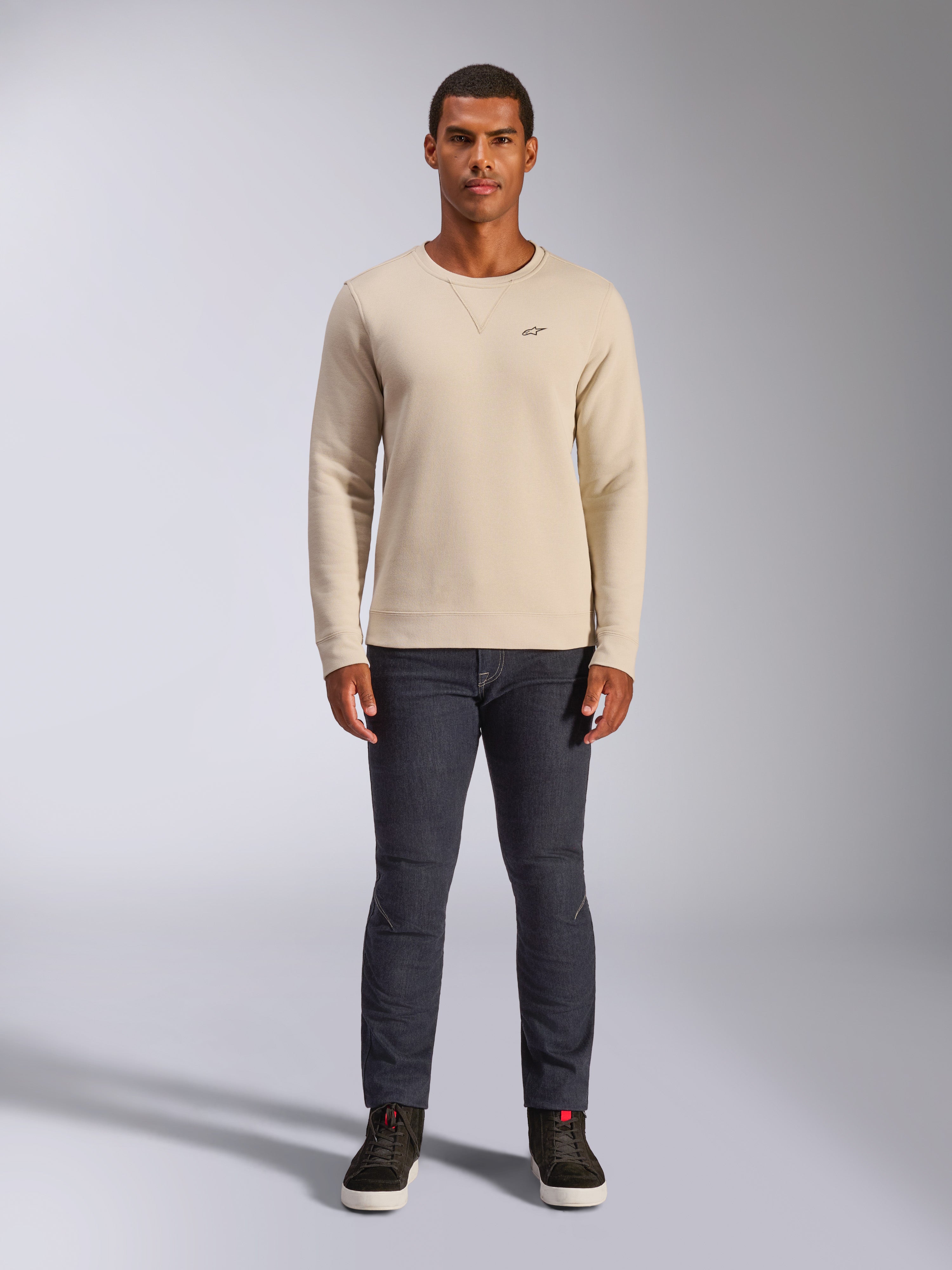 Alpinestars Positive Crew, Off-White Rundhals-Sweatshirt, kleines schwarzes Logo auf der Brust, gerippte Bündchen an Ärmeln und Saum, Dreiecksnaht am Ausschnitt, lässige Motorsport-Streetwear kombiniert mit dunkler Jeans
