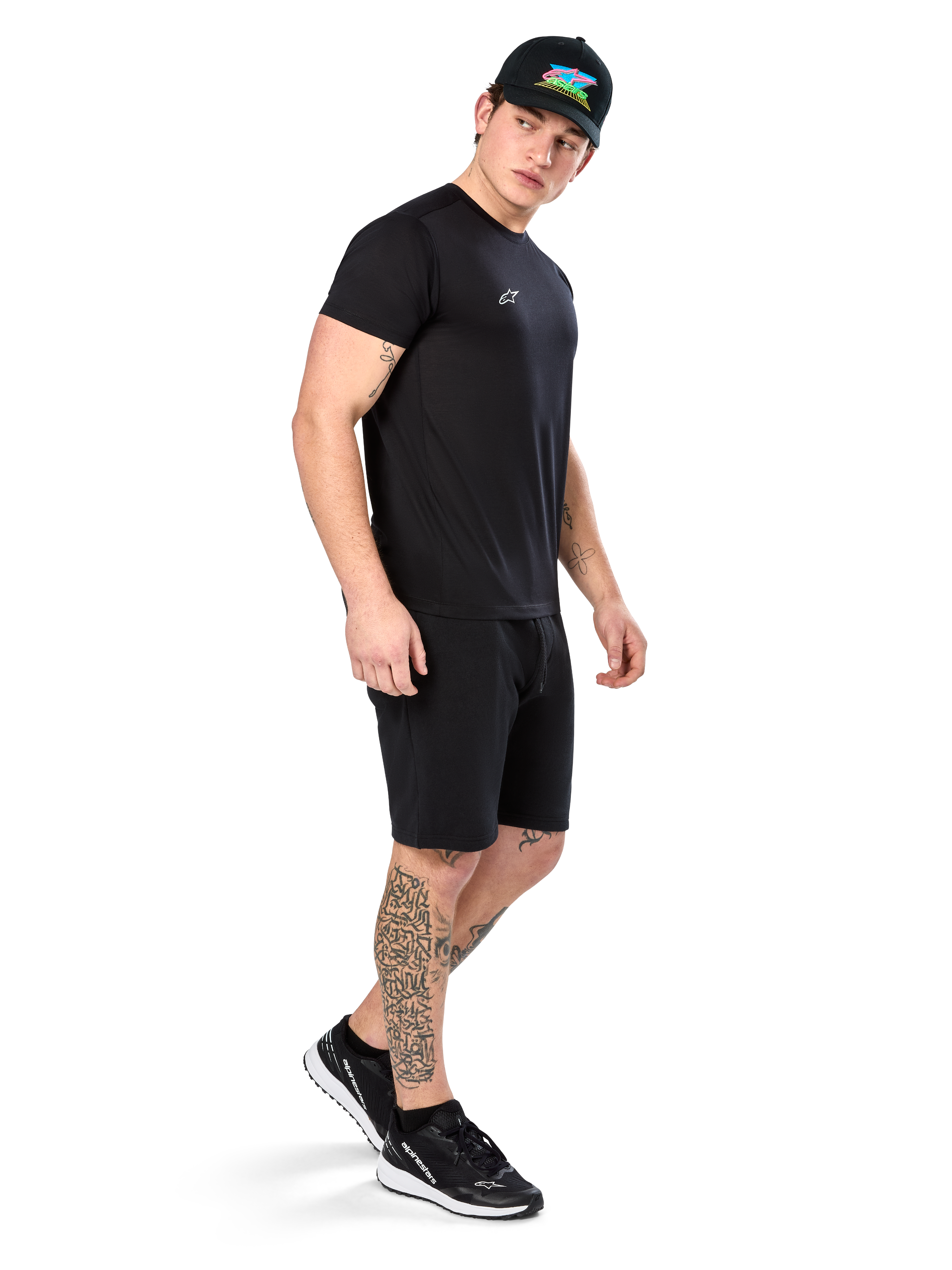 Man with tattoos wearing Alpinestars Point Performance Tee, Short sleeve black performance t-shirt, small white logo, paired with black shorts, a black cap with colorful graphic, and black and white sneakers, sporty casual lifestyle