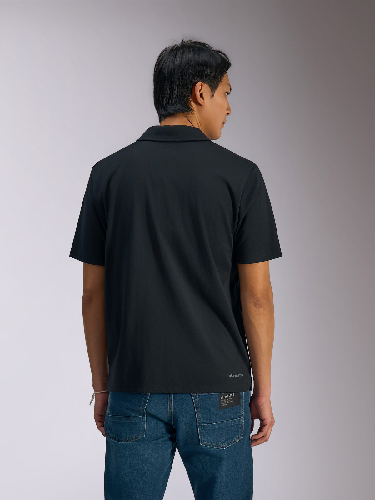 Alpinestars Impetus Sport Polo, schwarzes Kurzarm-Poloshirt, Rückansicht eines Mannes im Polo mit blauen Denim-Jeans, kleiner Alpinestars-Schriftzug am unteren Rücken, lässige vom Motorsport inspirierte Sportbekleidung