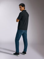 Alpinestars Impetus Sport Polo, Schwarz, kurzärmeliges Poloshirt mit Mock-Mesh-Gewebe und 3-Knopf-Leiste, getragen von einem Model mit blauen Jeans und schwarzen Sneakern, lässige vom Motorsport inspirierte Streetwear