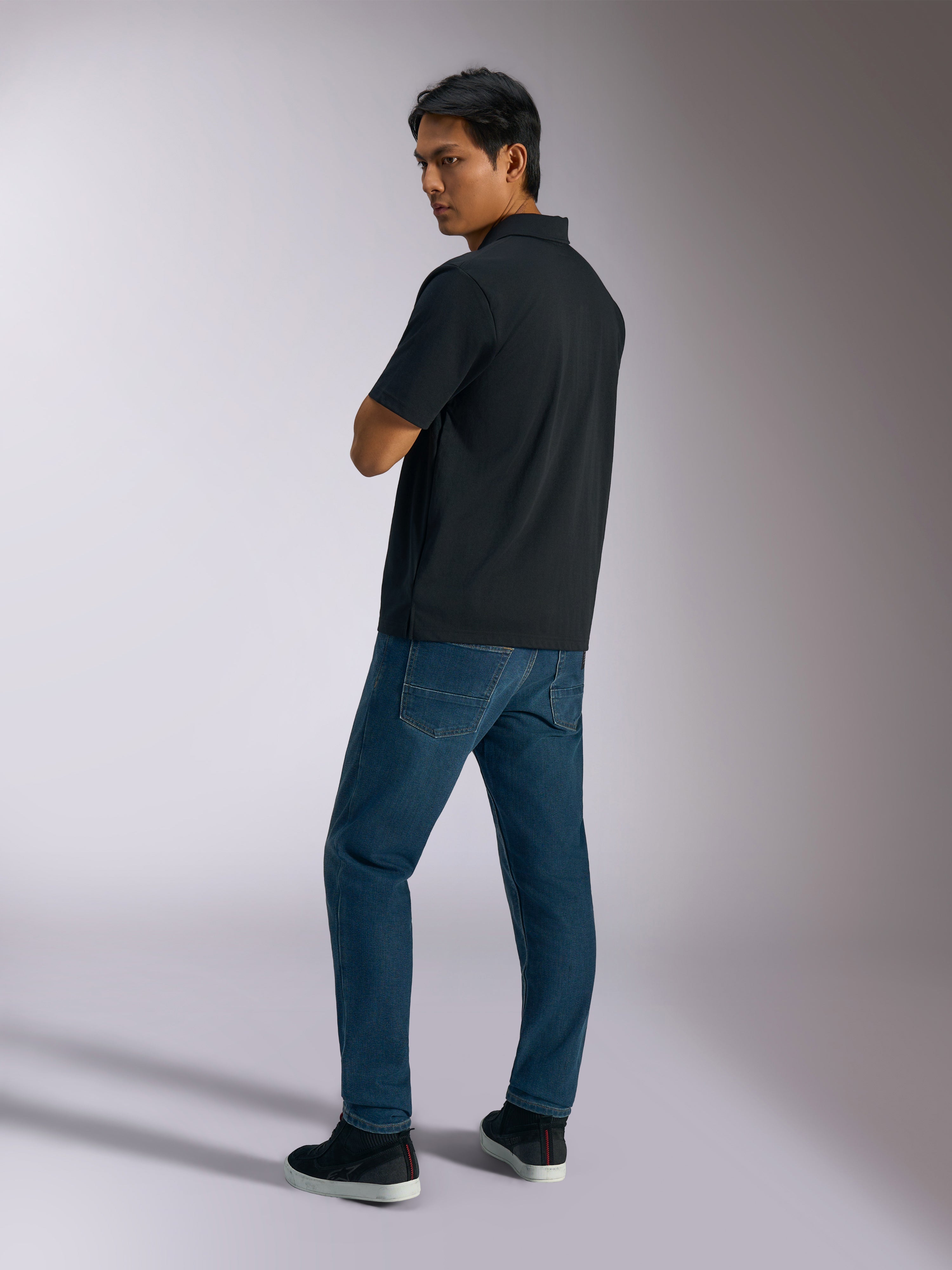 Alpinestars Impetus Sport Polo, Schwarz, kurzärmeliges Poloshirt mit Mock-Mesh-Gewebe und 3-Knopf-Leiste, getragen von einem Model mit blauen Jeans und schwarzen Sneakern, lässige vom Motorsport inspirierte Streetwear
