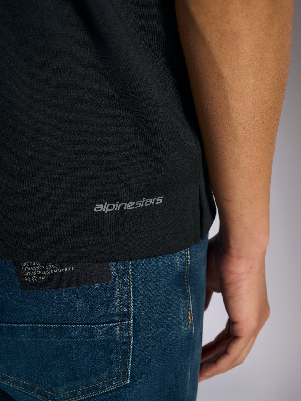 Alpinestars Impetus Sport Polo, Detailansicht des unteren Saums und Seitenschlitzes, Schwarz mit grauem Alpinestars-Schriftzug, lässige, vom Motorsport inspirierte Sportbekleidung