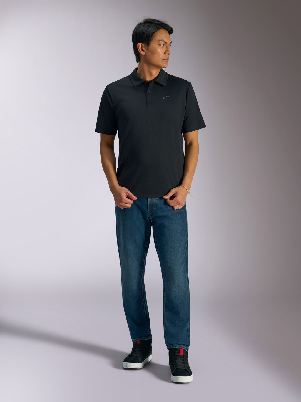 Alpinestars Impetus Sport Polo, schwarzes Kurzarm-Polohemd mit 3-Knopf-Leiste und grauem Astars-Logo auf der Brust, getragen von einem Mann mit blauen Jeans und schwarzen High-Top-Sneakern, lässige, vom Motorsport inspirierte Sportbekleidung