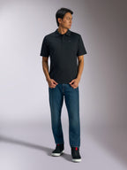 Alpinestars Impetus Sport Polo, schwarzes Kurzarm-Polohemd mit 3-Knopf-Leiste und grauem Astars-Logo auf der Brust, getragen von einem Mann mit blauen Jeans und schwarzen High-Top-Sneakern, lässige, vom Motorsport inspirierte Sportbekleidung