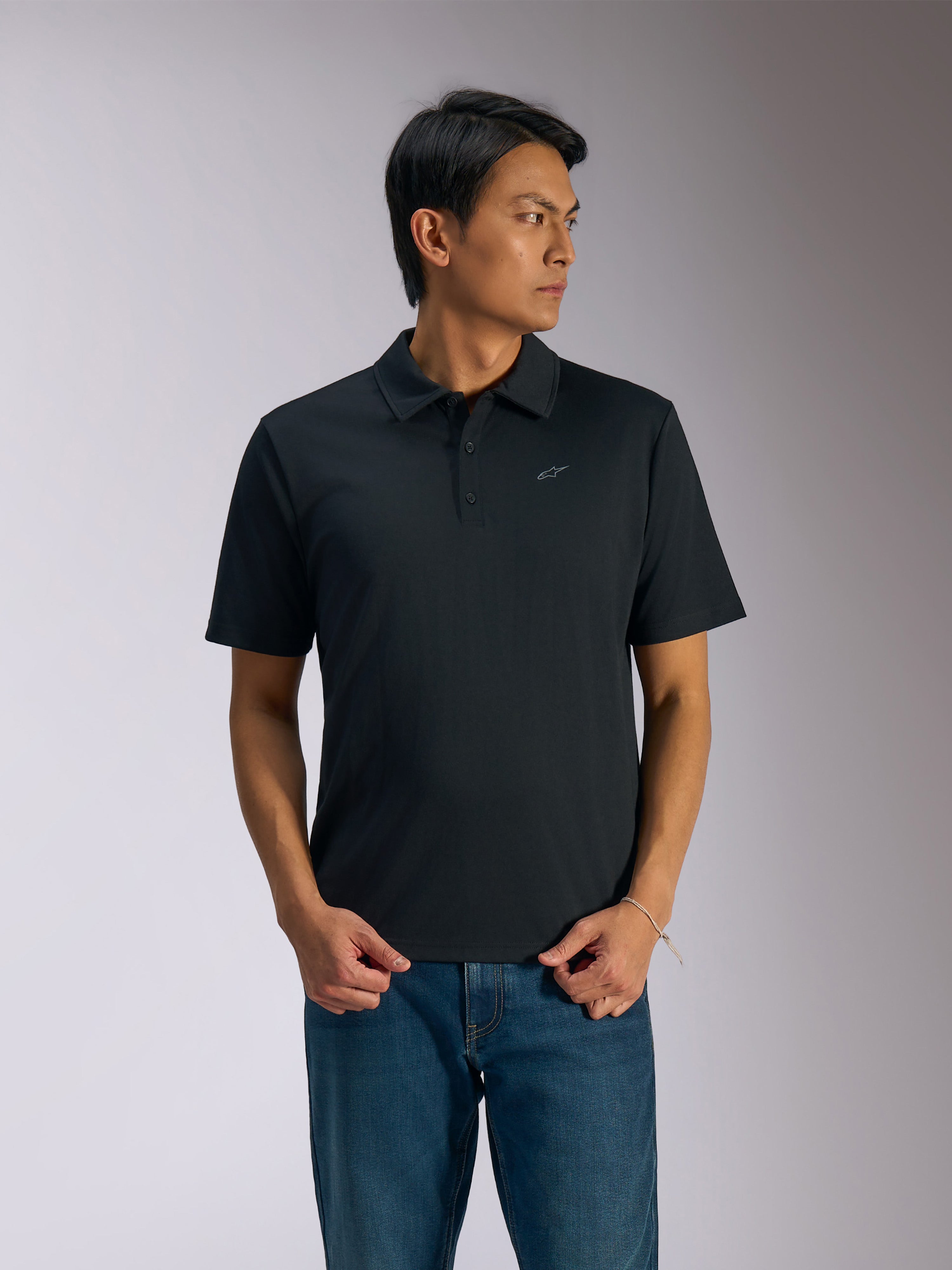 Alpinestars Impetus Sport Polo, schwarzes Kurzarm-Poloshirt, Drei-Knopf-Leiste und Selbstkragen, kleines graues Astars-Logo auf der linken Brust, getragen mit blauen Jeans, lässige, vom Motorsport inspirierte Sportbekleidung