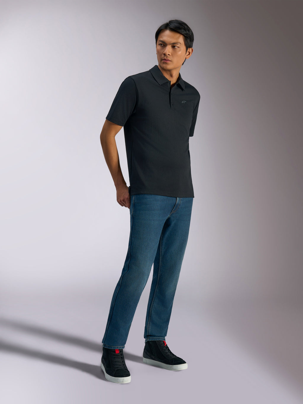 Alpinestars Impetus Sport Polo, Kurzarm-Poloshirt, Schwarz, mit dezentem Brustlogo und Drei-Knopf-Leiste, getragen von einem Modell mit blauen Jeans und schwarzen Sneakern, lässige, vom Motorsport inspirierte Lifestyle-Bekleidung