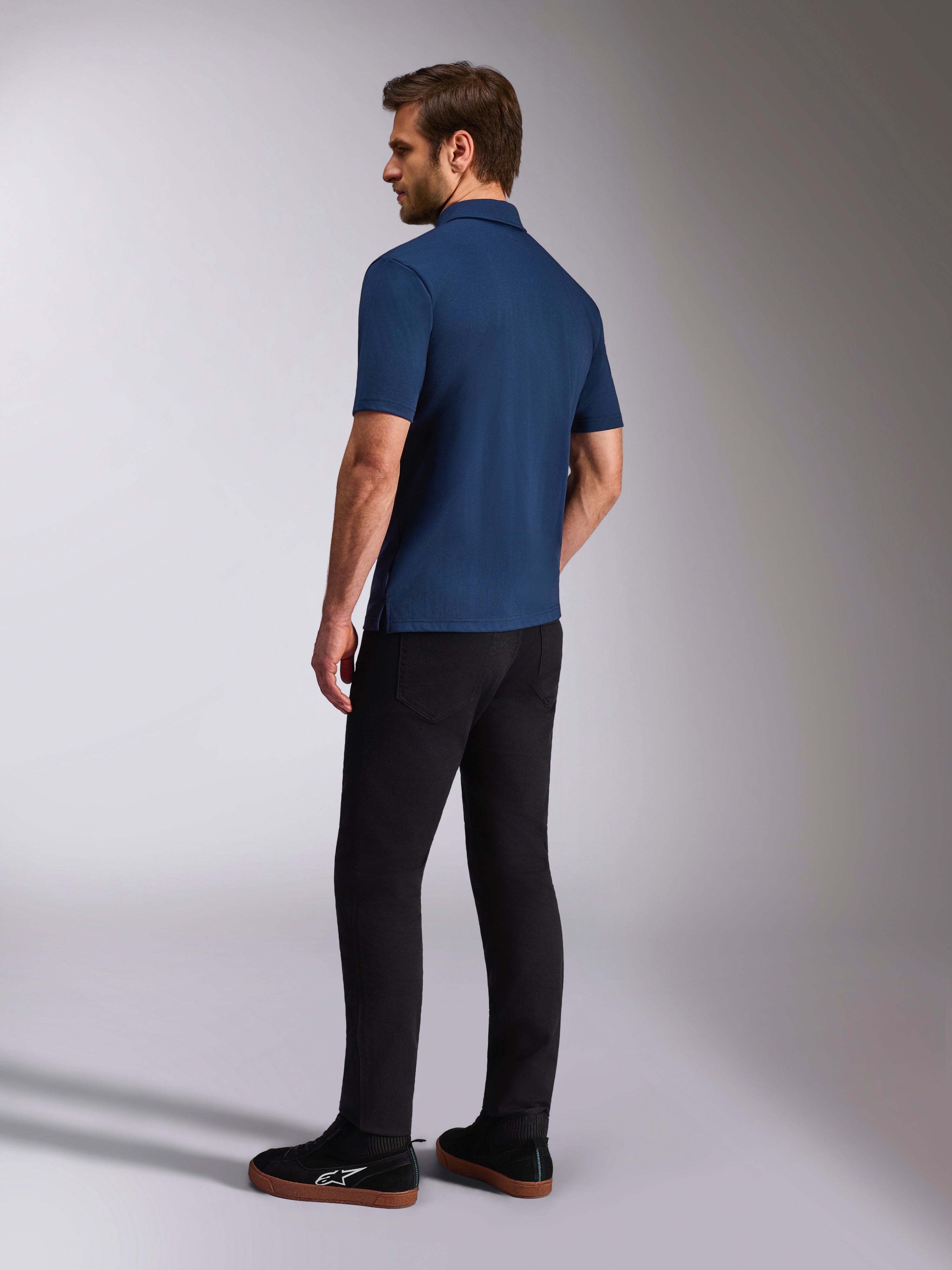 Alpinestars Impetus Sport Polo, kurzärmeliges Poloshirt in Marineblau, Rück- und Seitenansicht am Model, kombiniert mit schwarzer Hose und schwarzen Sneakern, lässige Motorsport-Lifestyle-Sportbekleidung