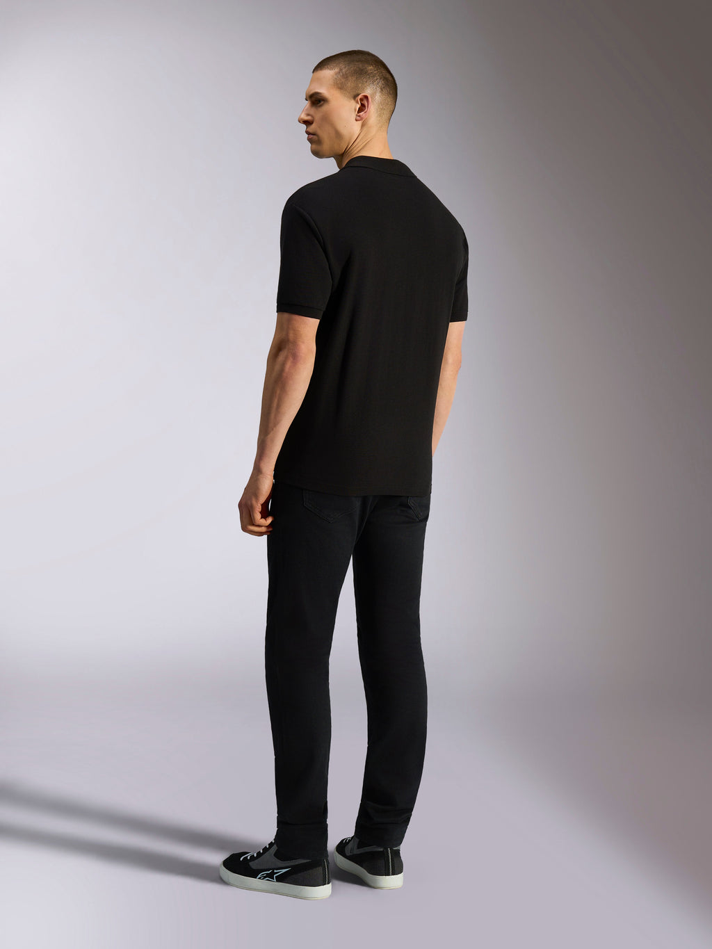 Alpinestars Endeavor Classic Polo, kurzärmeliges Piqué-Poloshirt, Schwarz, Rückansicht eines männlichen Models in schwarzer Hose und Sneakern, lässige motorsportinspirierte Streetwear
