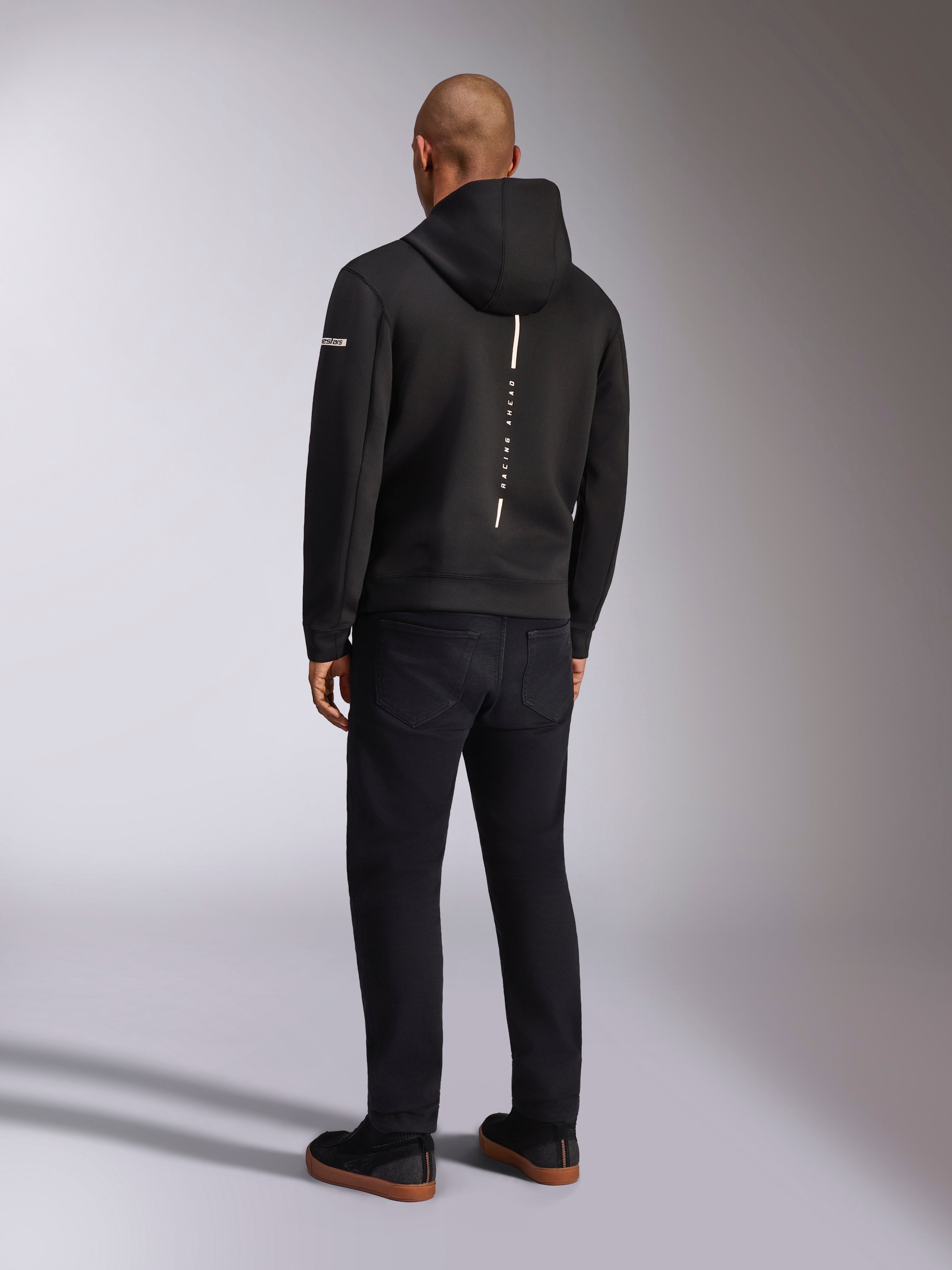 Alpinestars Insight Hoodie, schwarzer Sport-Kapuzenpullover, Rückansicht mit vertikalem weißem Streifen und „RACING AHEAD“-Schriftzug, Ärmellogo, kombiniert mit schwarzer Hose und Sneakern, lässiges, vom Motorsport inspiriertes Design