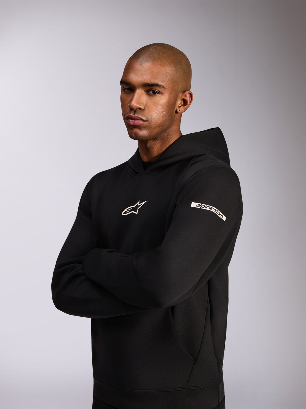 Alpinestars Insight Hoodie, lässiger Sportswear-Kapuzenpullover, Schwarz, weißes Astars-Logo auf der Brust und Wortlogo am Oberärmel, Model trägt Kapuzenpullover mit verschränkten Armen, hochwertige Doppelvlies-Konstruktion
