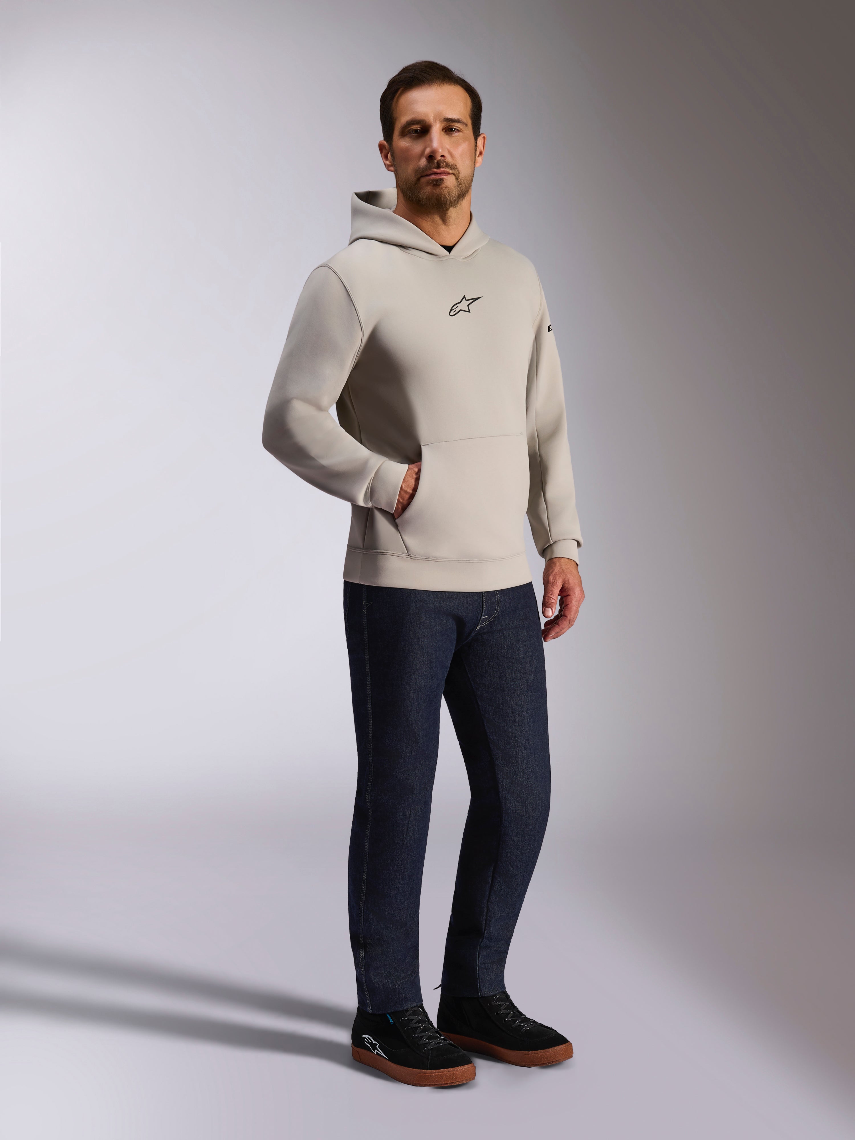 Alpinestars Insight Kapuzenpullover, lässige Streetwear, Zementgrau mit schwarzem Astars-Logo auf der Brust, Pullover-Design mit Kängurutasche, getragen mit dunkelblauer Jeans und schwarzen Sneakern, moderner, vom Motorsport inspirierter Stil