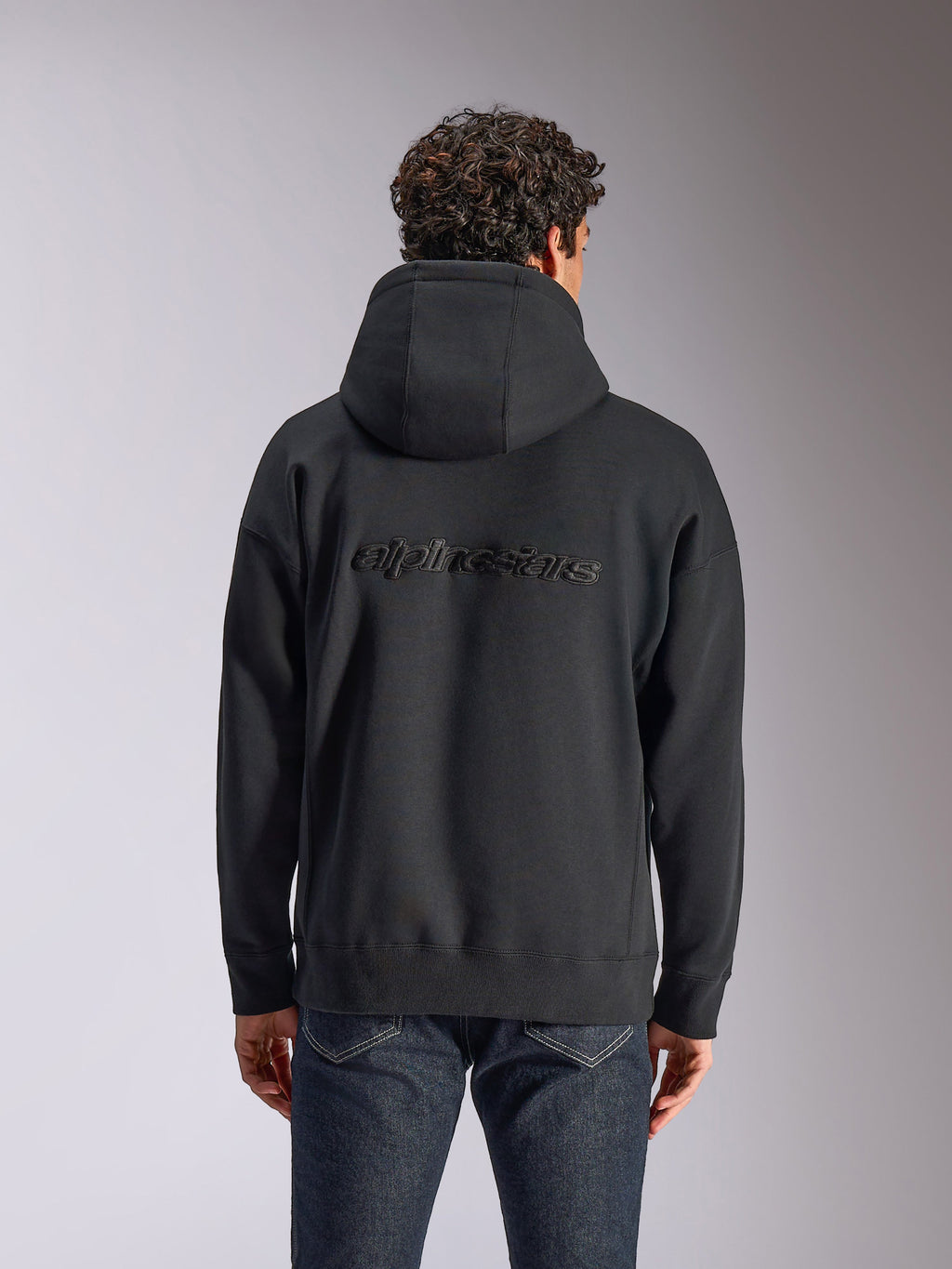 Alpinestars Emergent Oversized Hoodie, Rückansicht eines schwarzen Kapuzenpullovers an einem Model, besticktes Alpinestars-Wortlogo auf dem Rücken, Oversized-Passform, lässige Streetwear kombiniert mit dunkler Jeans