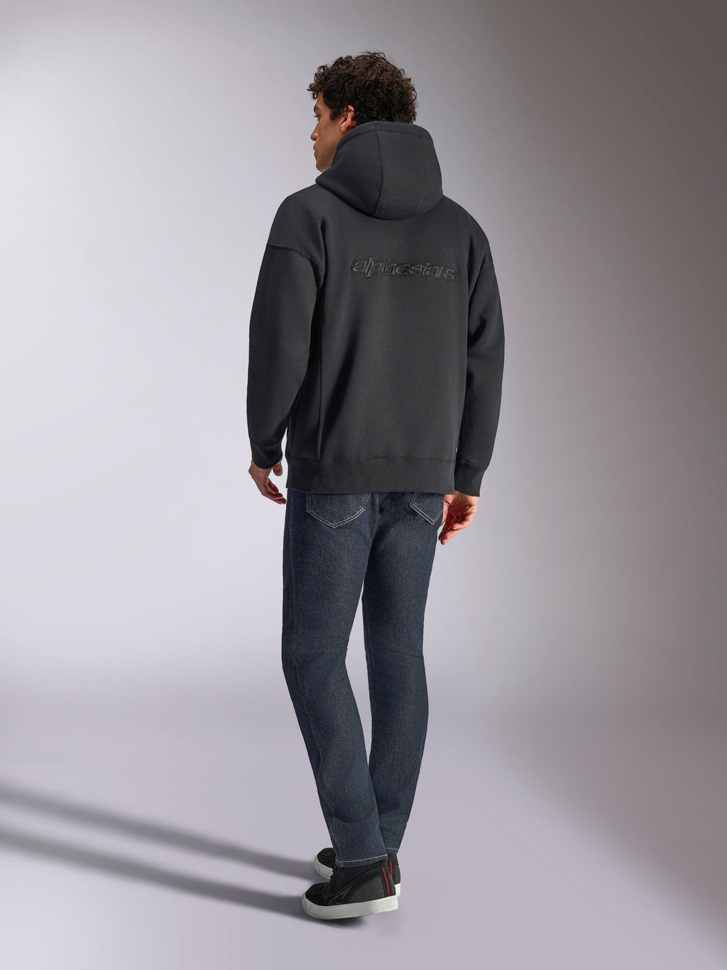 Alpinestars Emergent Oversized Hoodie, Schwarz, Rückansicht mit 3D-gesticktem Alpinestars-Wortlogo über den Schultern, Oversize-Passform, kombiniert mit dunkelblauer Denim-Jeans und schwarzen Sneakern, lässige, vom Motorsport inspirierte Streetwear