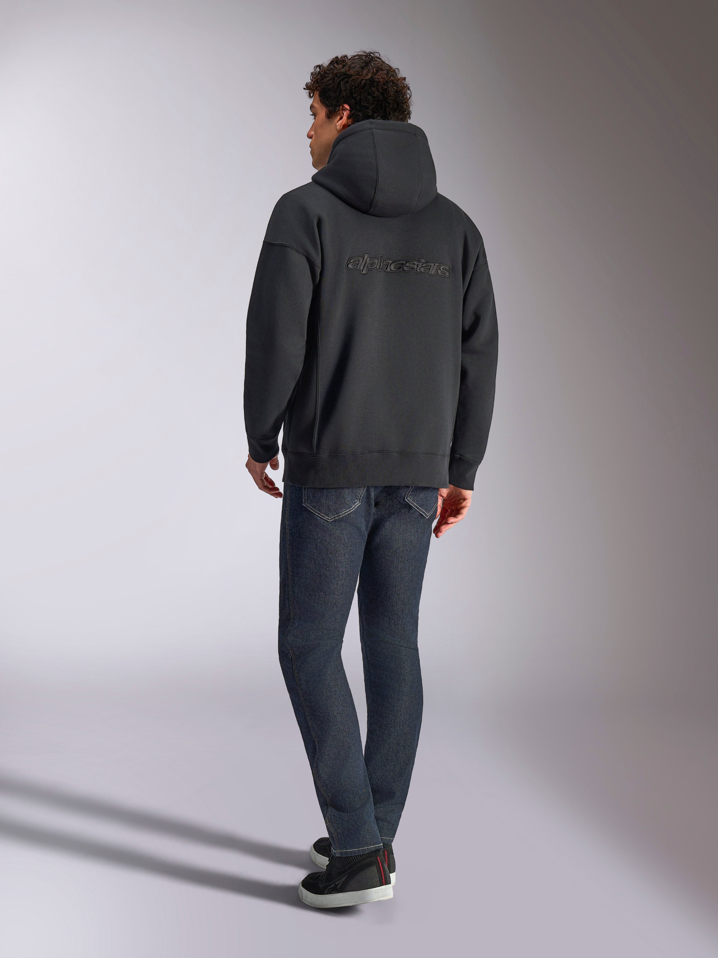 Alpinestars Emergent Oversized Hoodie, Schwarz, Rückansicht mit 3D-gesticktem Alpinestars-Wortlogo über den Schultern, Oversize-Passform, kombiniert mit dunkelblauer Denim-Jeans und schwarzen Sneakern, lässige, vom Motorsport inspirierte Streetwear