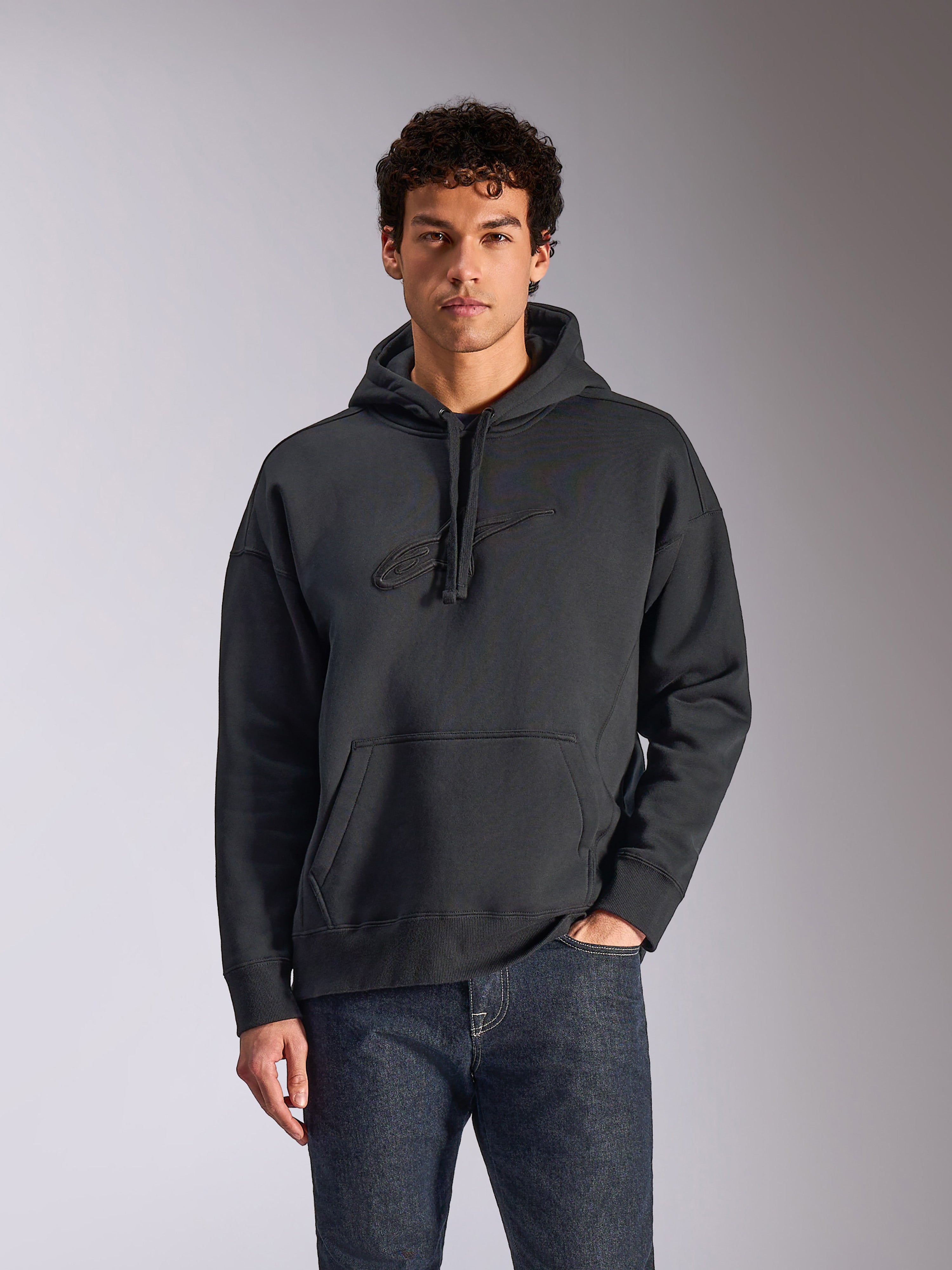 Alpinestars Emergent Oversized Hoodie, Schwarz, männliches Model trägt einen Oversized-Fleece-Kapuzenpullover, tonales 3D-gesticktes Astars-Logo, Kängurutasche, Kapuze mit Kordelzug, lässige vom Motorsport inspirierte Streetwear