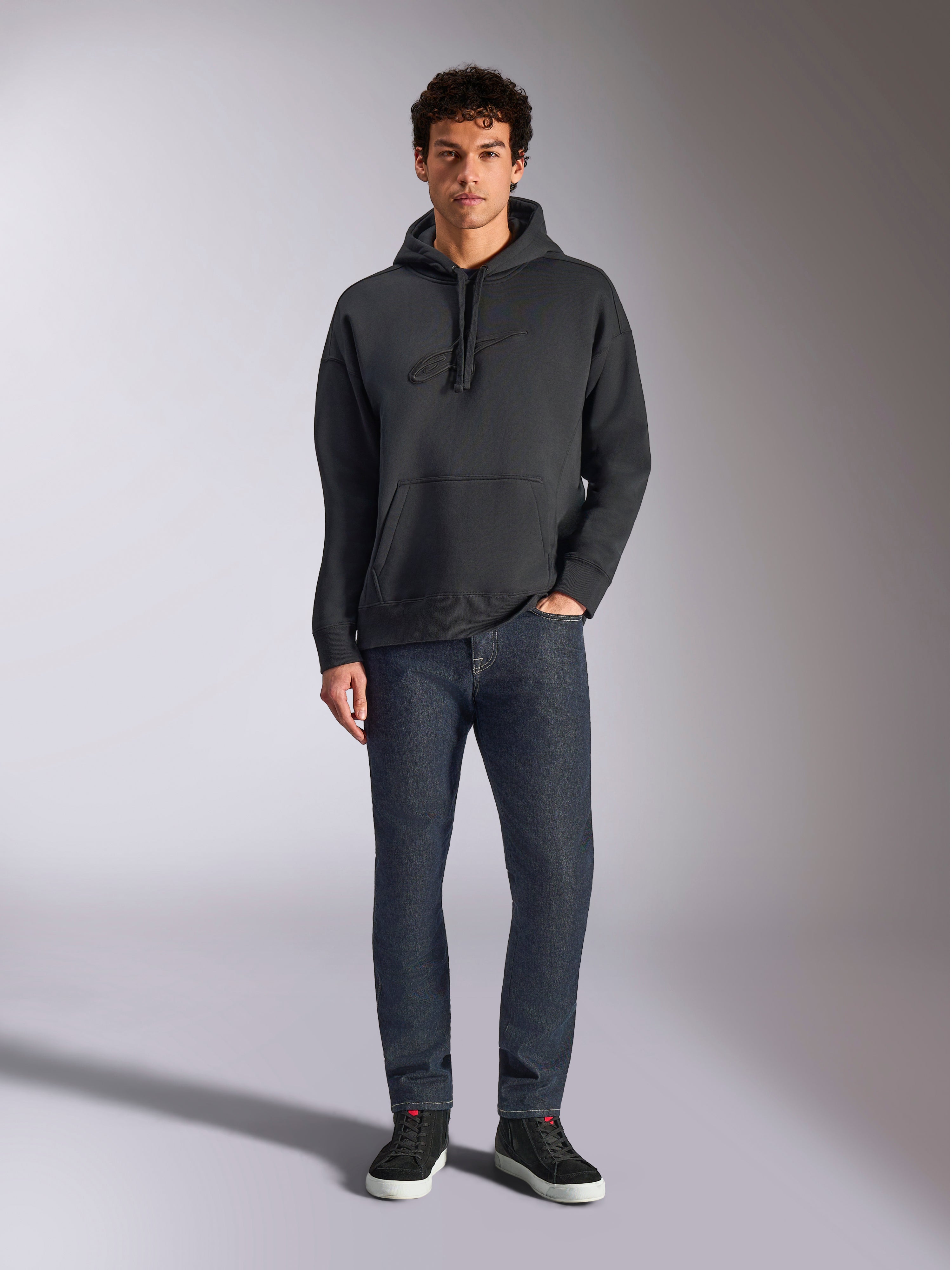 Alpinestars Emergent Oversized Hoodie, lässige Lifestyle-Streetwear, Schwarz mit tonalem 3D-Sticklogo, Kapuze mit Kordelzug, Kängurutasche, Oversized-Passform, getragen von einem Model mit dunkelblauen Jeans und schwarzen Sneakern