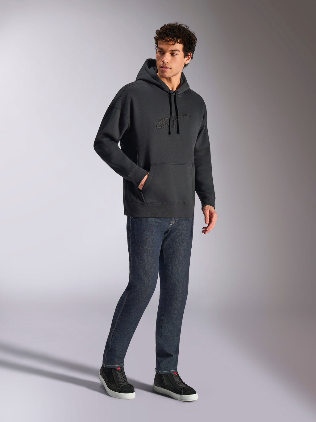 Alpinestars Emergent Oversized Hoodie, schwarze lässige Streetwear, tonales 3D-besticktes Astars-Logo auf der Brust, Kängurutasche, Oversized-Passform, getragen von einem männlichen Model mit dunkelblauen Jeans und schwarzen Sneakern