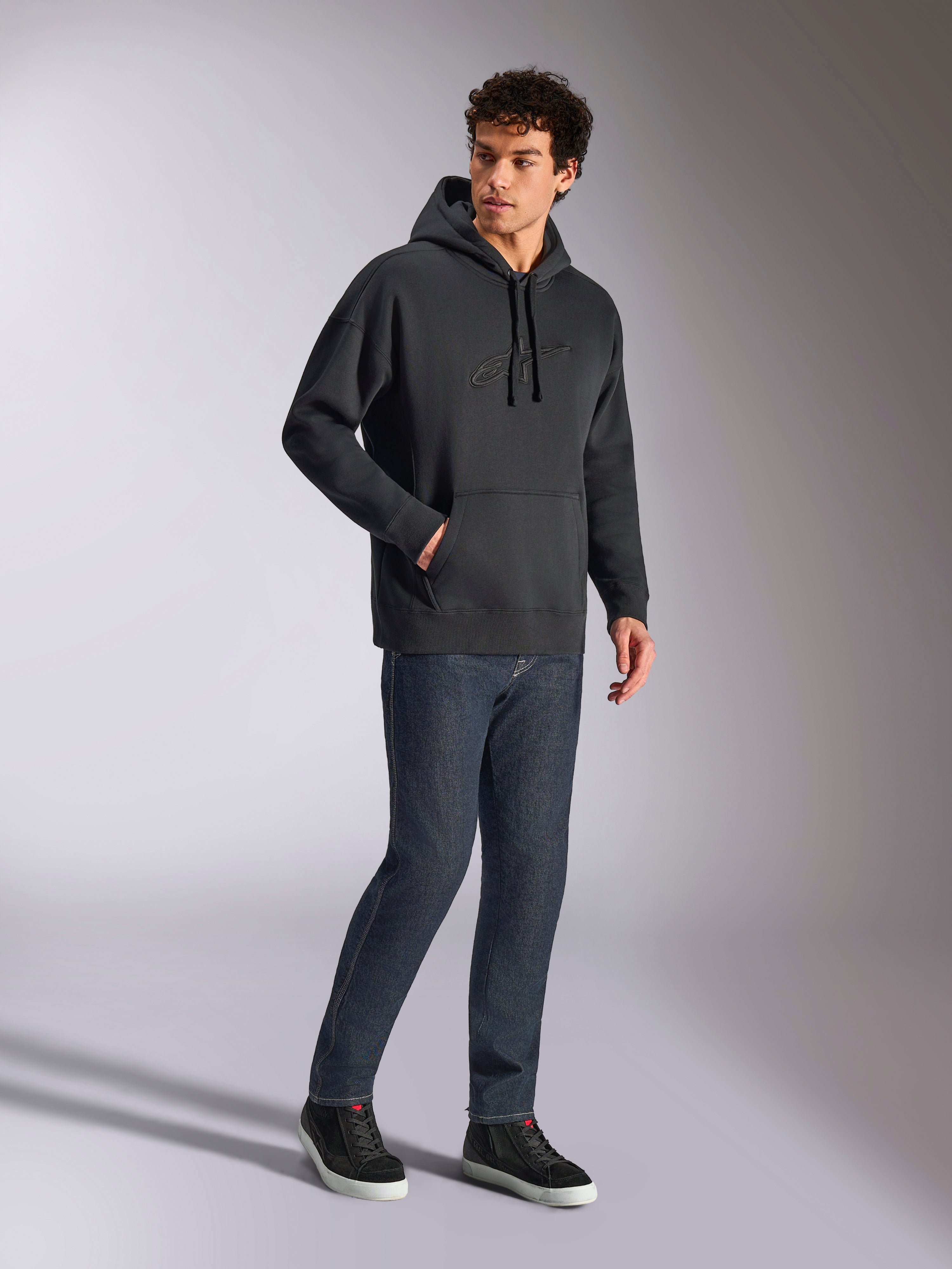 Alpinestars Emergent Oversized Hoodie, schwarze lässige Streetwear, tonales 3D-besticktes Astars-Logo auf der Brust, Kängurutasche, Oversized-Passform, getragen von einem männlichen Model mit dunkelblauen Jeans und schwarzen Sneakern