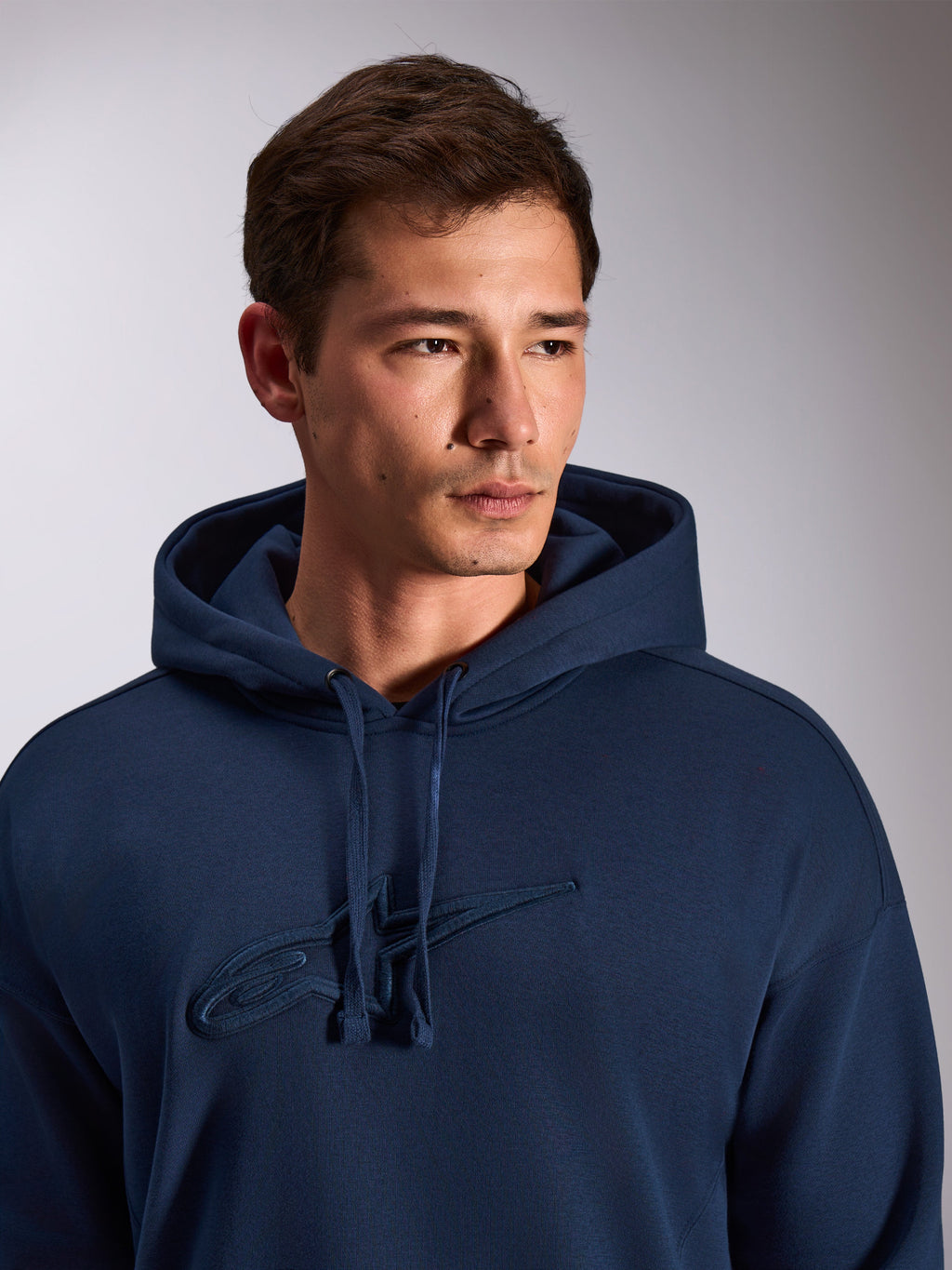 Alpinestars Emergent Oversized Hoodie, navyblau, Nahaufnahme eines Models, das einen Fleece-Kapuzenpullover trägt, tonale 3D-Stickerei des Astars-Logos auf der Brust, verstellbare Kapuze mit Kordelzug, lässige Motorsport-Streetwear