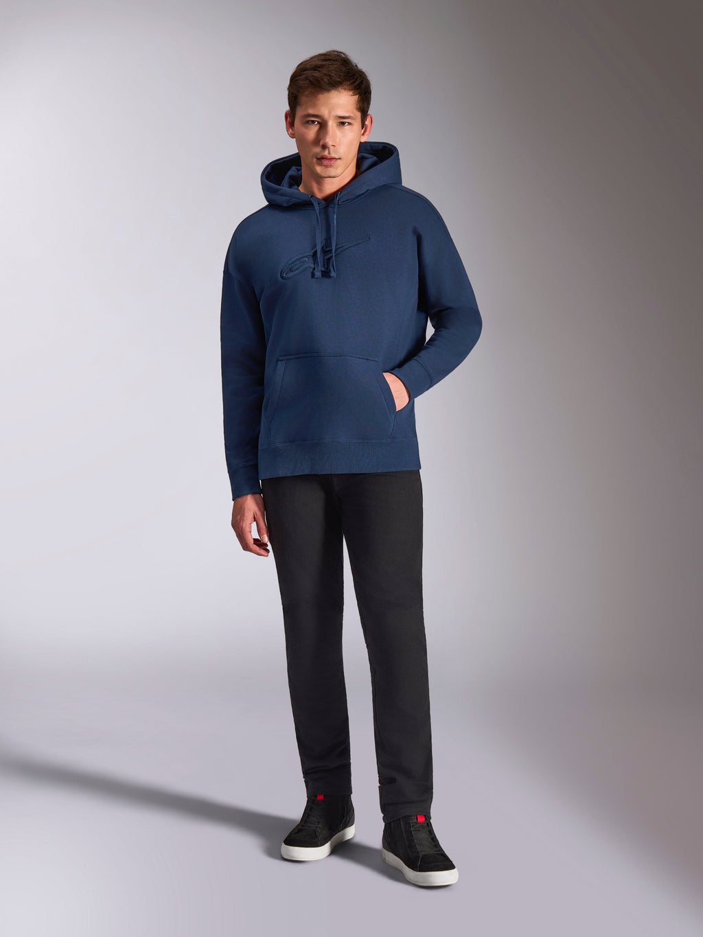 Alpinestars Emergent Oversized Hoodie, marineblauer Fleece-Kapuzenpullover, farblich abgestimmte 3D-Logostickerei auf der Vorderseite, getragen von einem Modell mit schwarzer Hose und Sneakern, lässiger Streetwear-Stil.